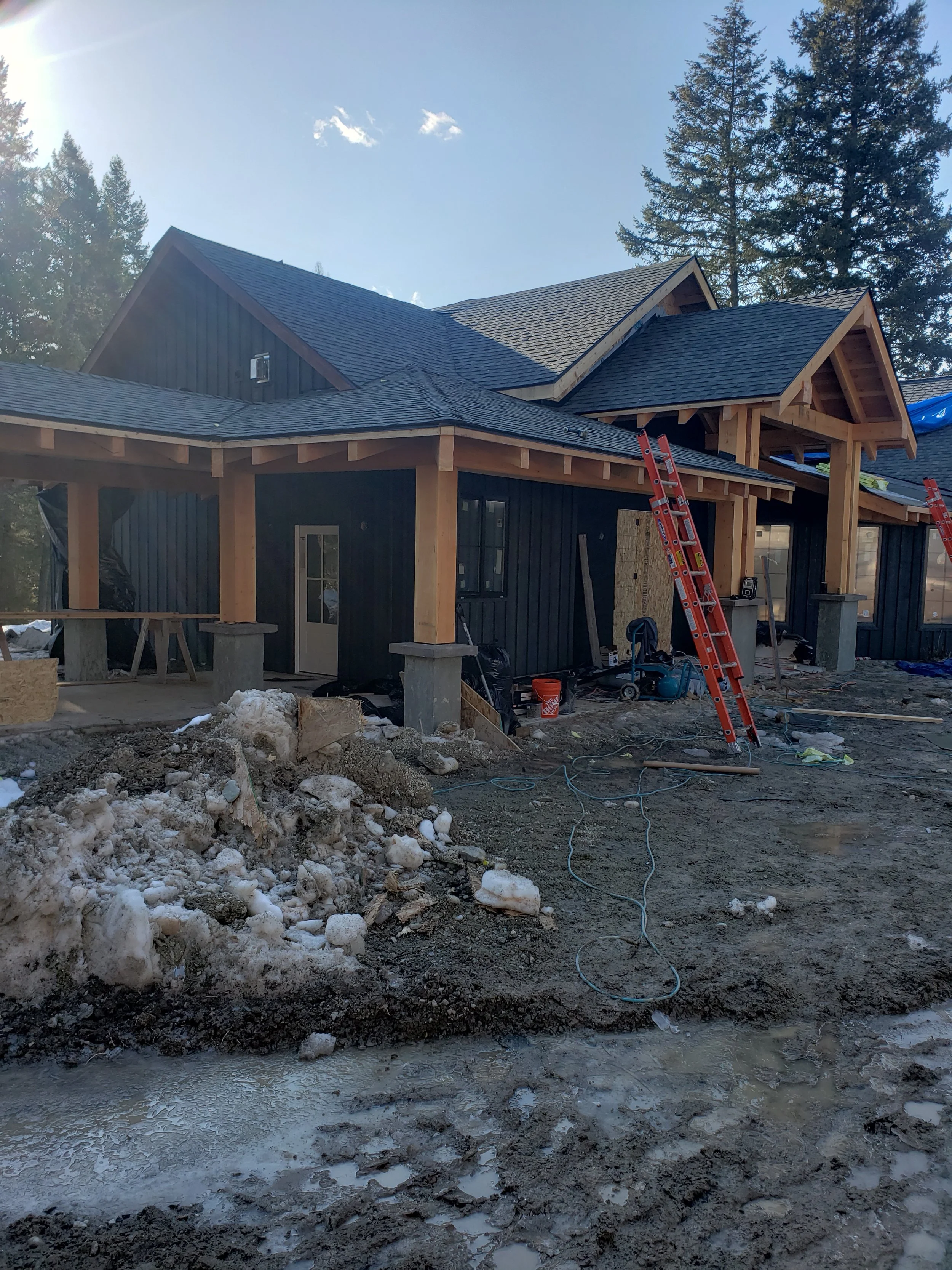 Under construction house with black exterior siding, wooden framing, and multiple roofing levels, surrounded by construction debris, ladders, and tall trees in the background.