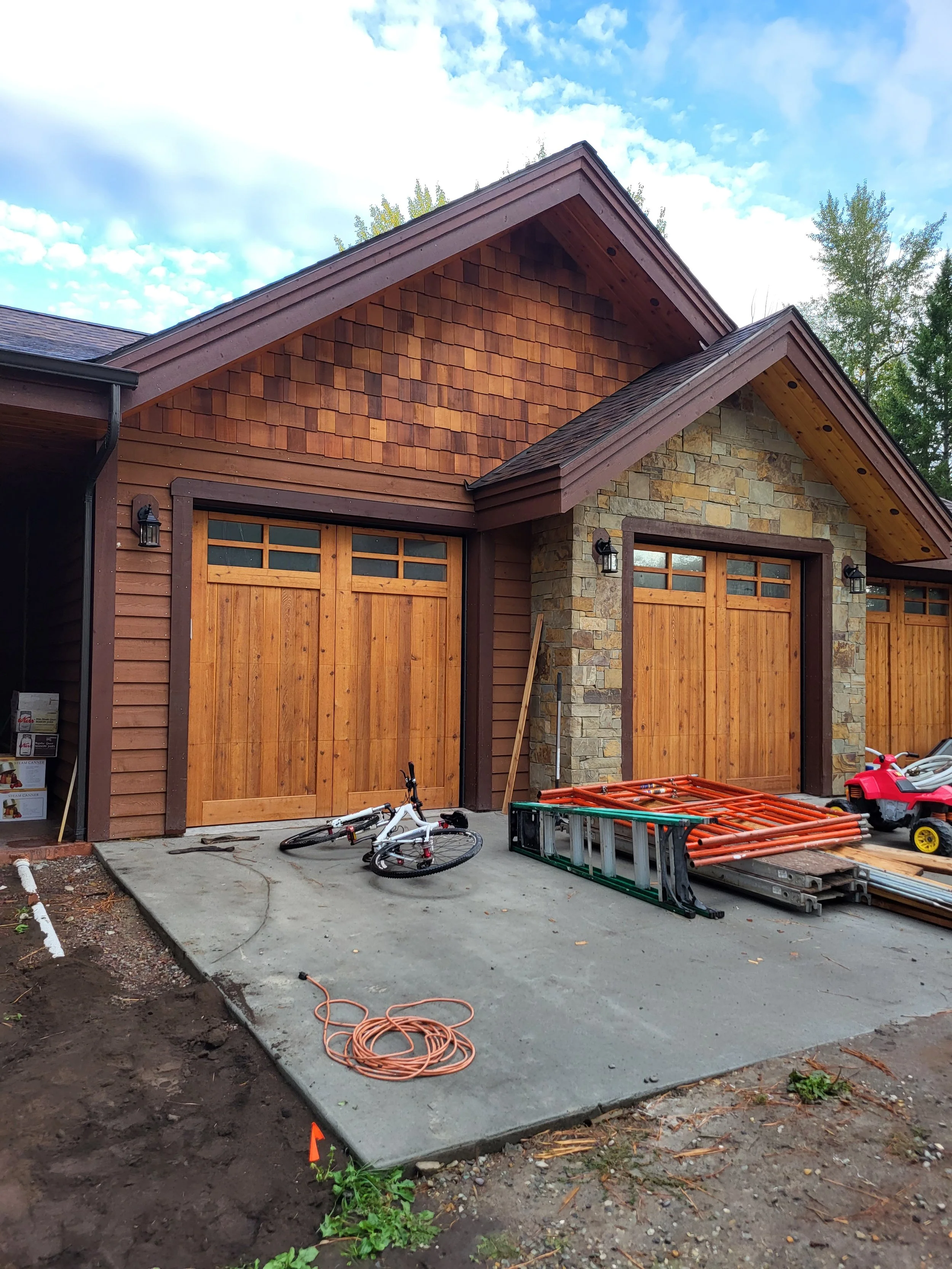 Construction site of a house with a concrete driveway, wooden garage doors, a bicycle, construction materials, and tools, with a partly cloudy sky in the background.