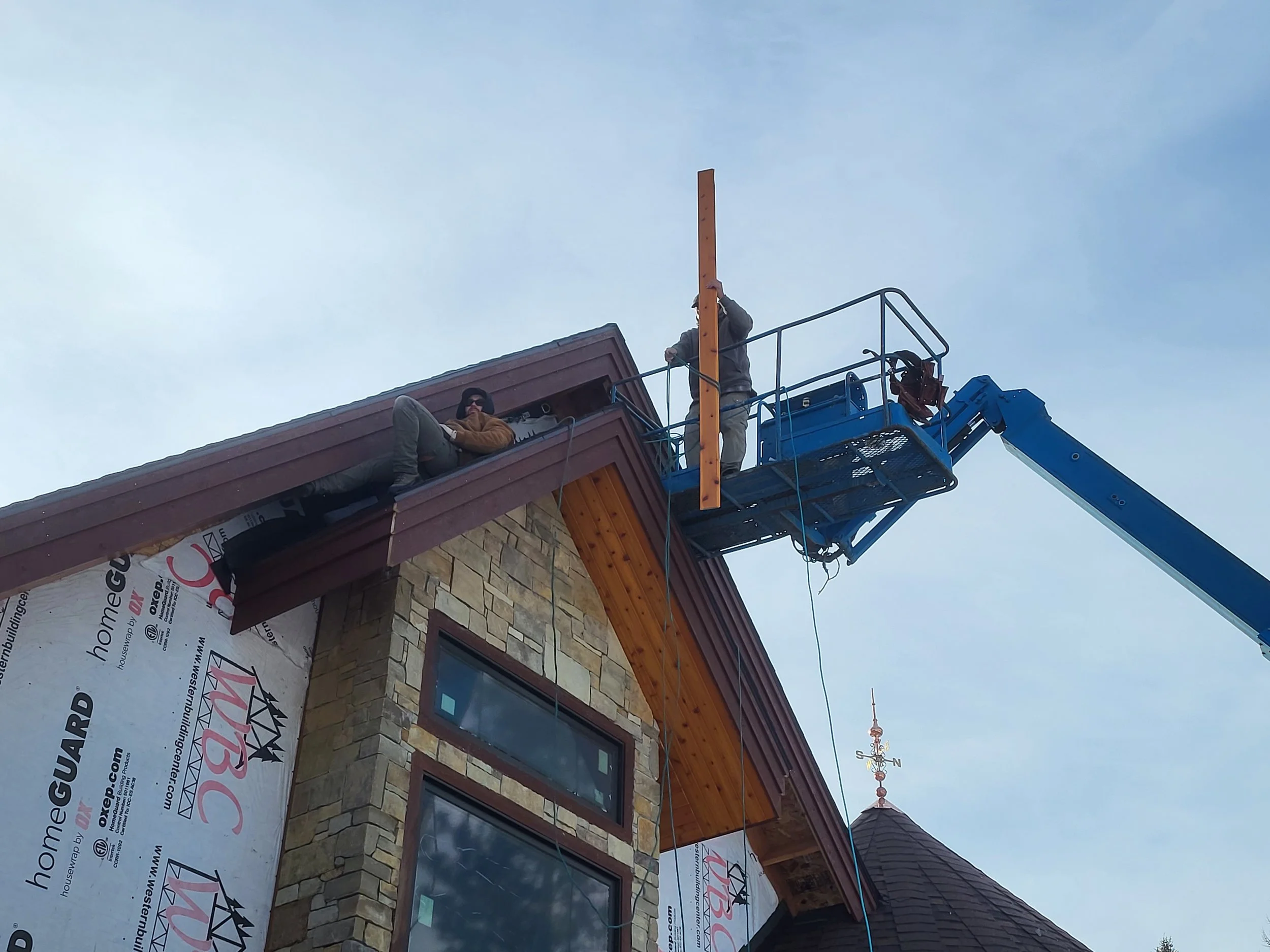 Two construction workers on a platform installing a vertical wooden piece on a house roof under cloudy sky.