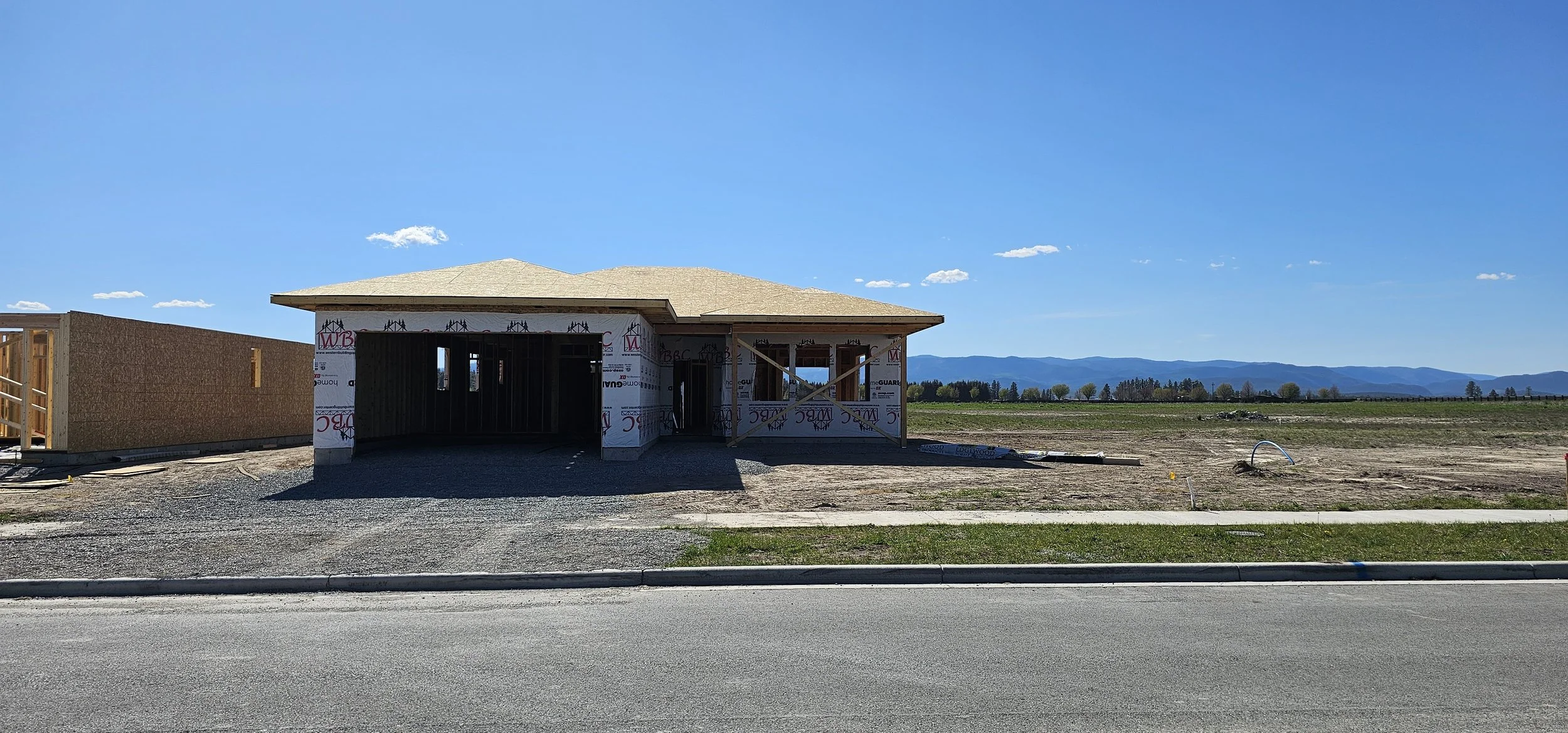 A house under construction in a rural area with mountains in the background, with unfinished walls and a partially built roof.