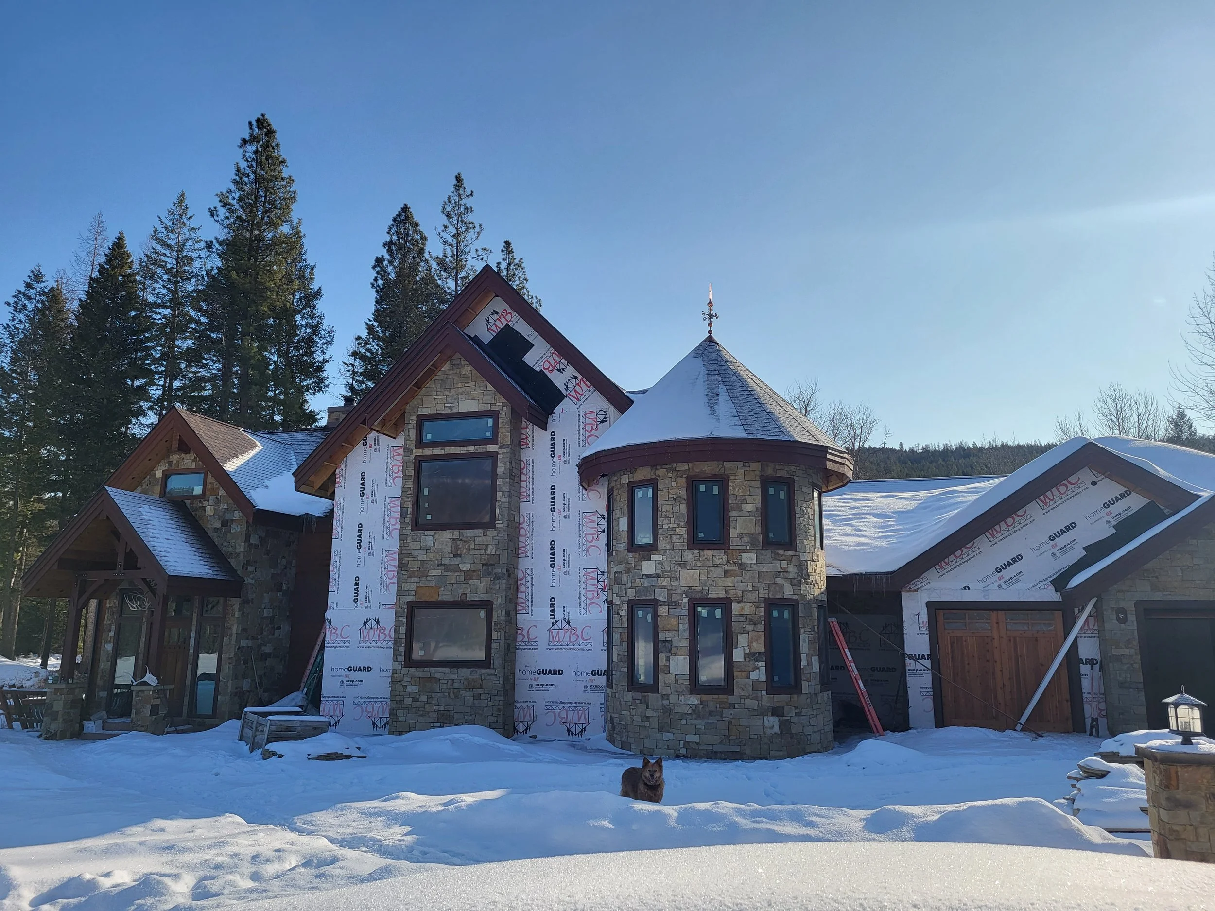 A house under construction with snow in the front yard, surrounded by trees and a clear blue sky.