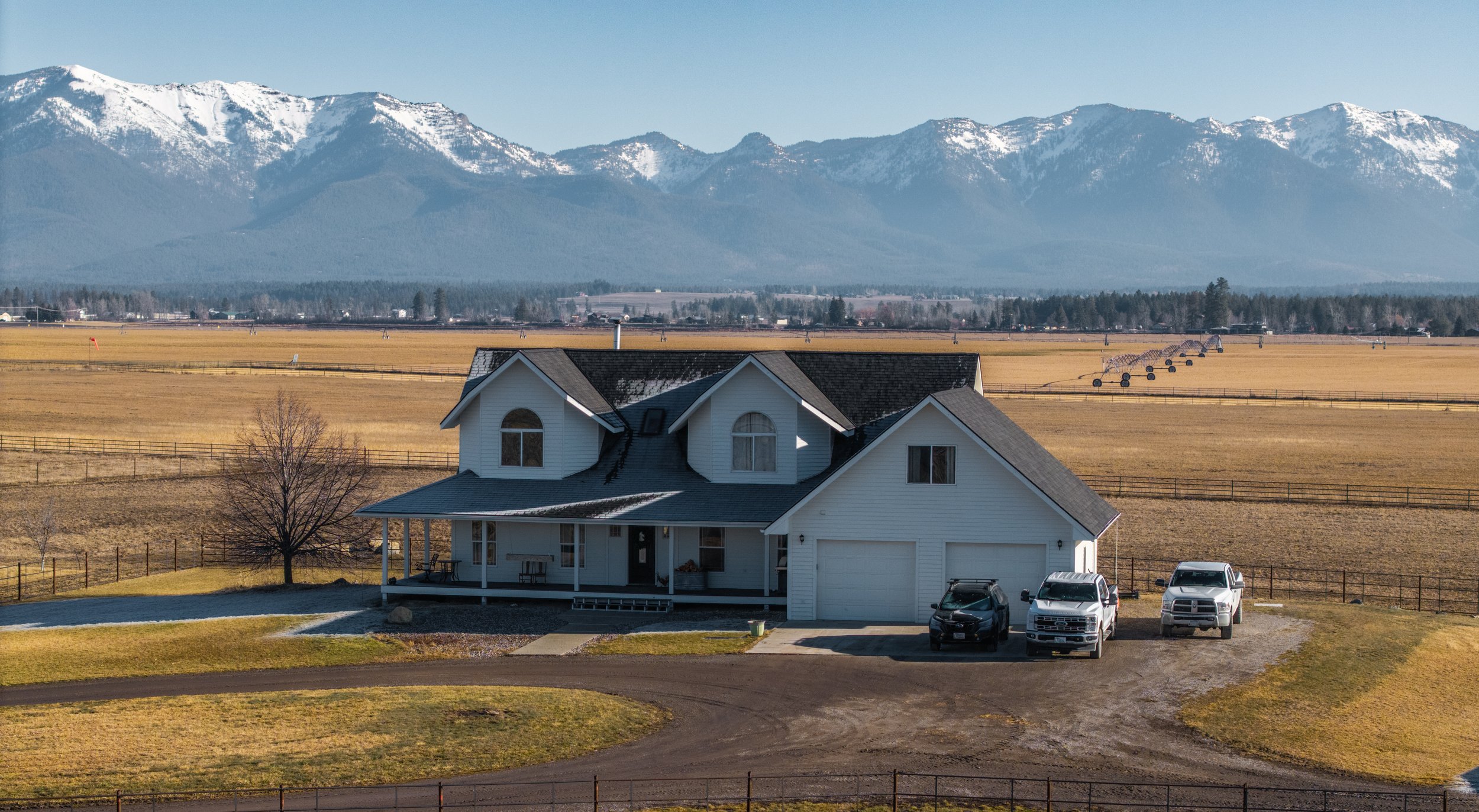 A white two-story house with a front porch and a detached garage, three vehicles parked in front, surrounded by a rural landscape with fields, mountains in the background, and snow on the mountain peaks.