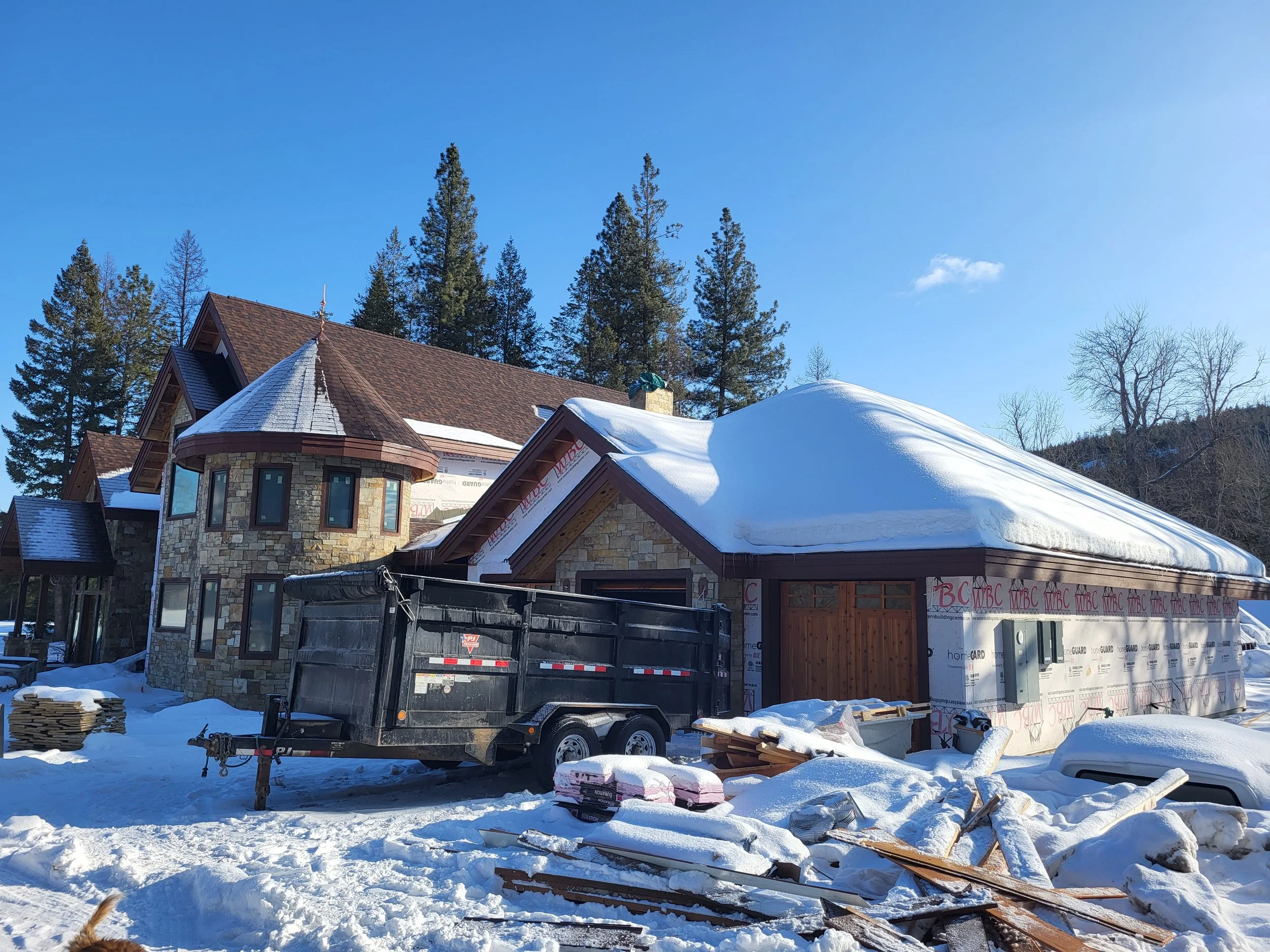 A house under construction covered in snow, with a black trailer in front, surrounded by construction materials and debris on snowy ground, with trees and a blue sky in the background.