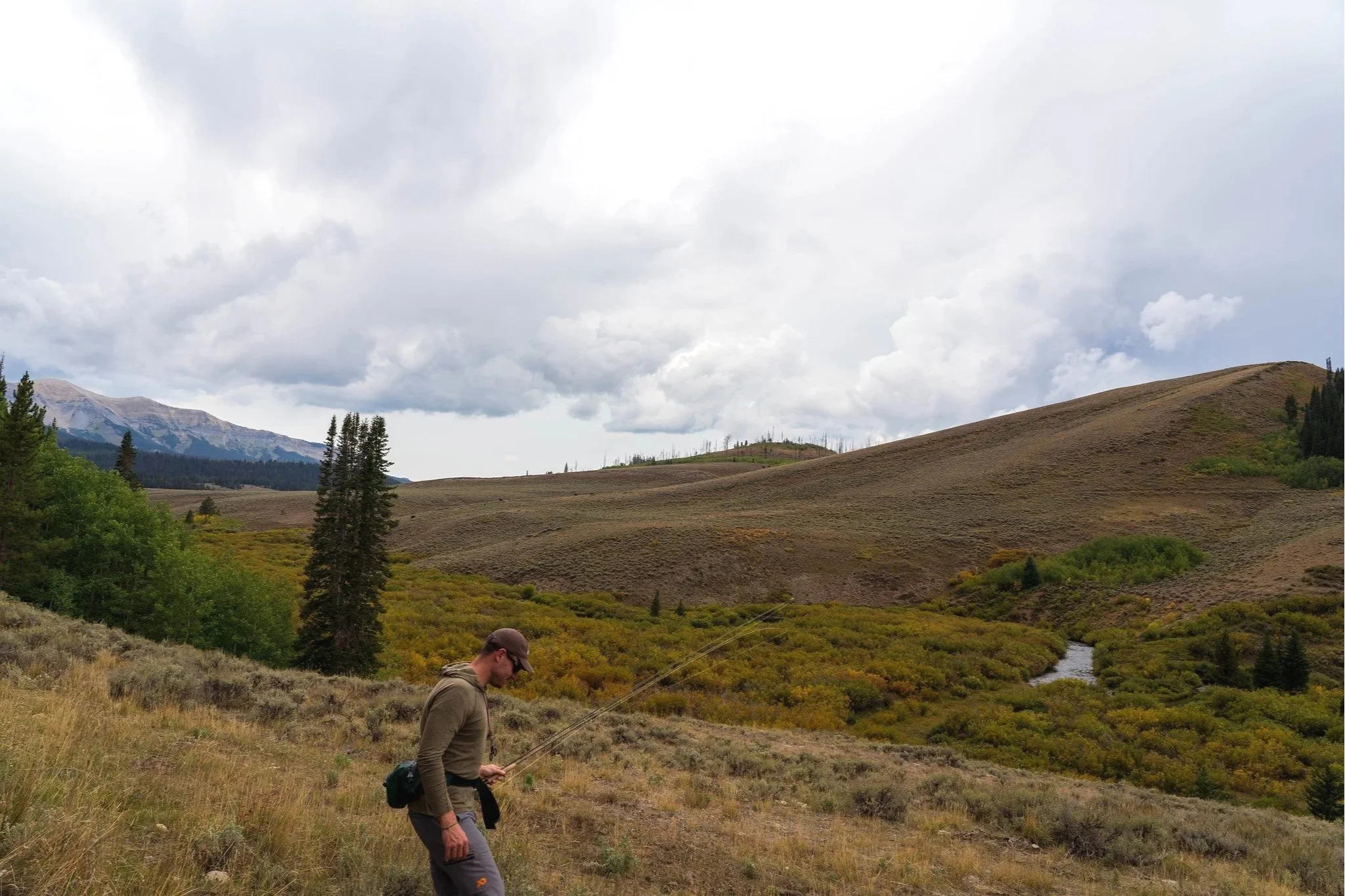 A fisherman walks toward a small stream in a meadow below a mountain range.