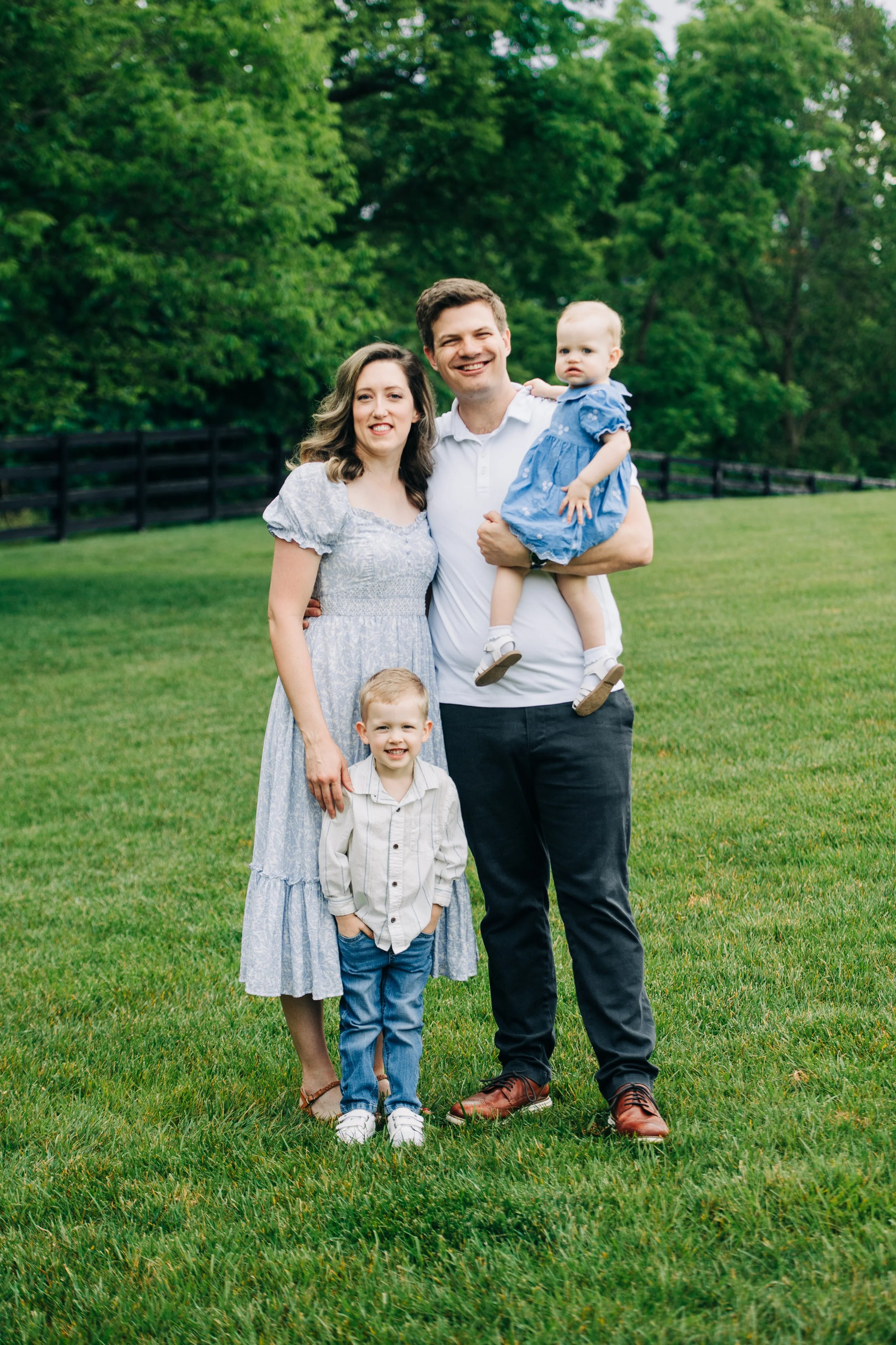 A happy family of four standing on a green lawn with trees and a black wooden fence in the background.