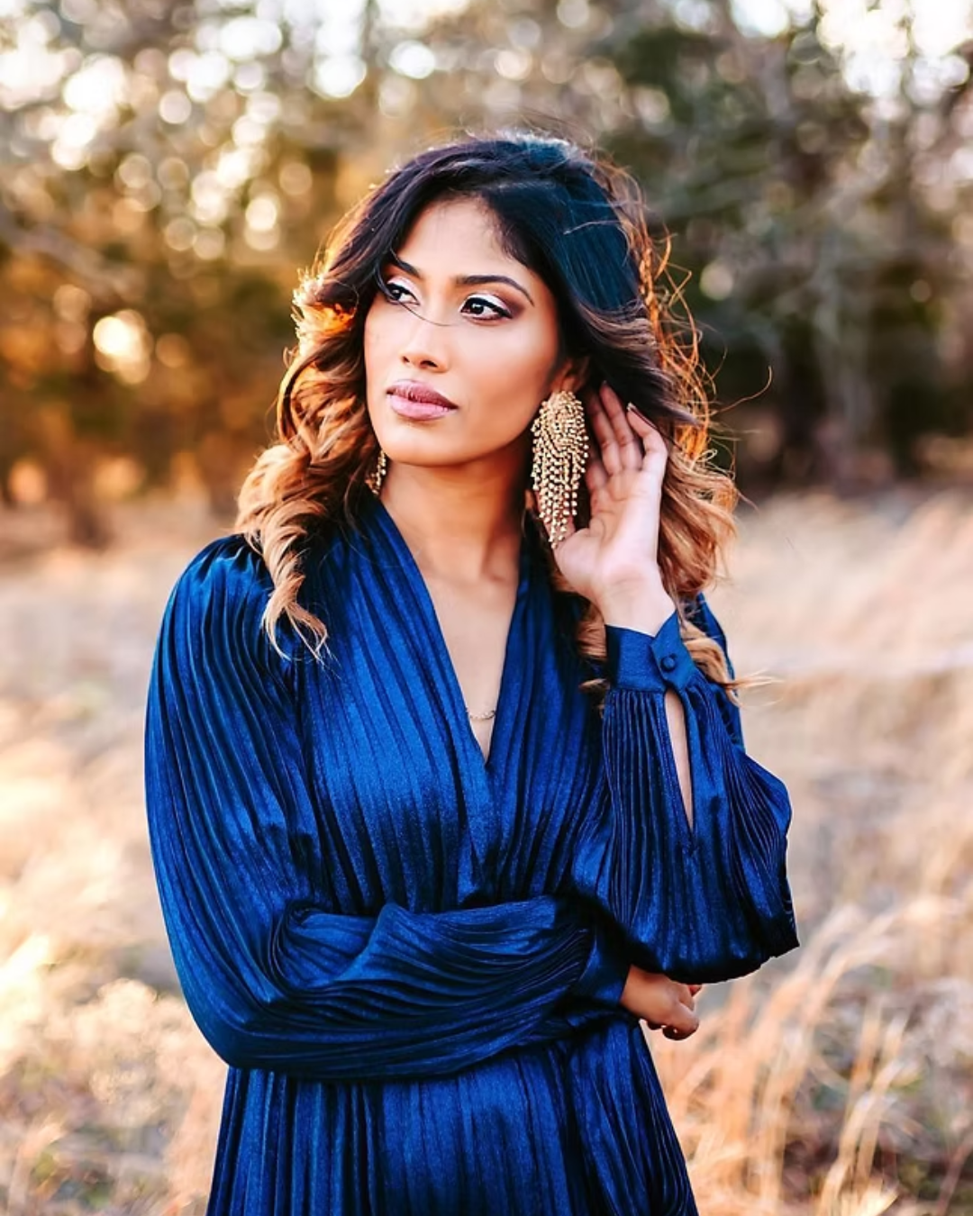 A woman with long, wavy hair wearing large, gold earrings and a deep blue dress, standing outdoors with a blurred background of trees and sunlight.