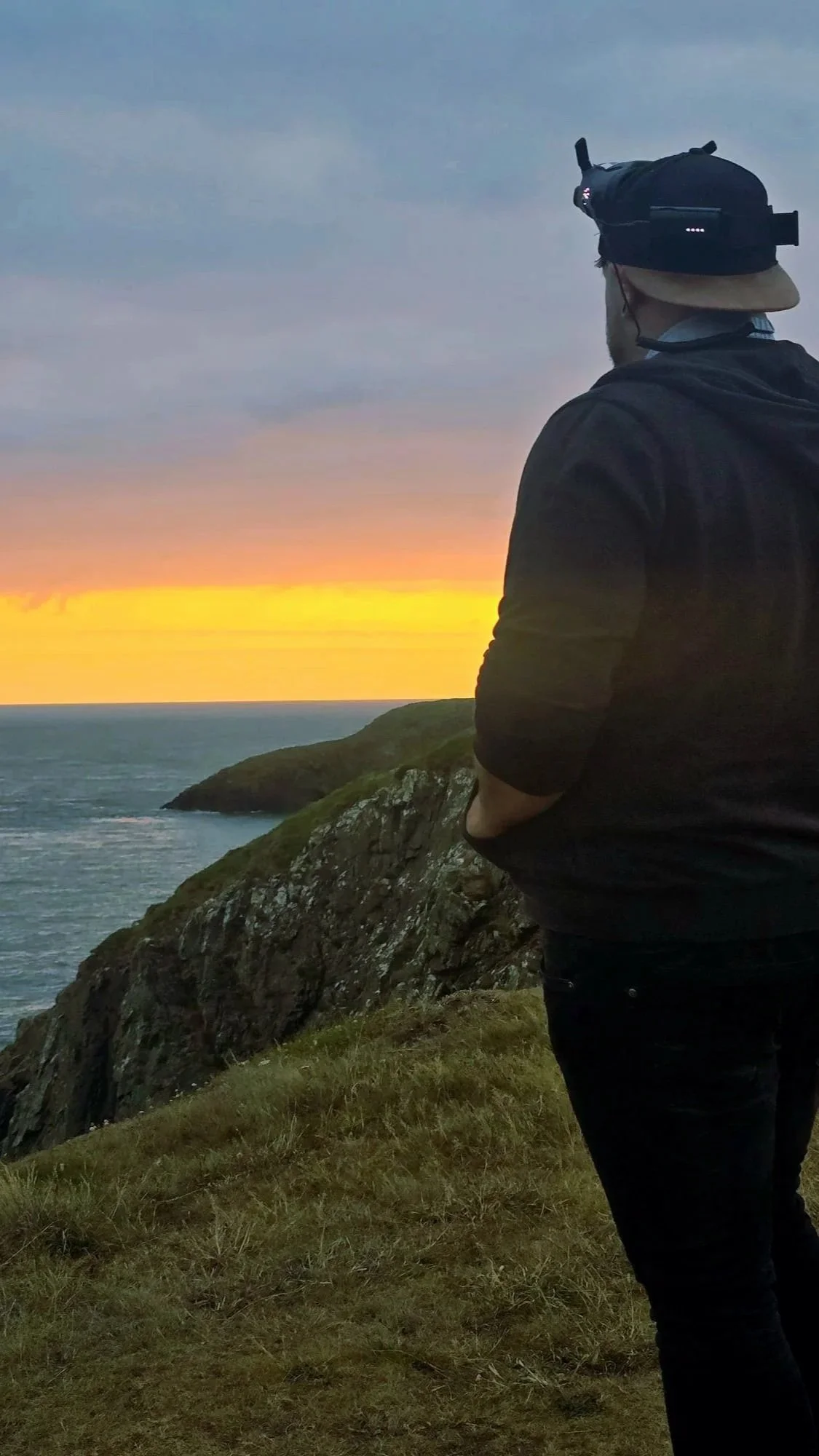 Man standing on grassy cliff overlooking the ocean at sunset, wearing a black jacket, black pants, and a black cap with a camera mounted on his head.