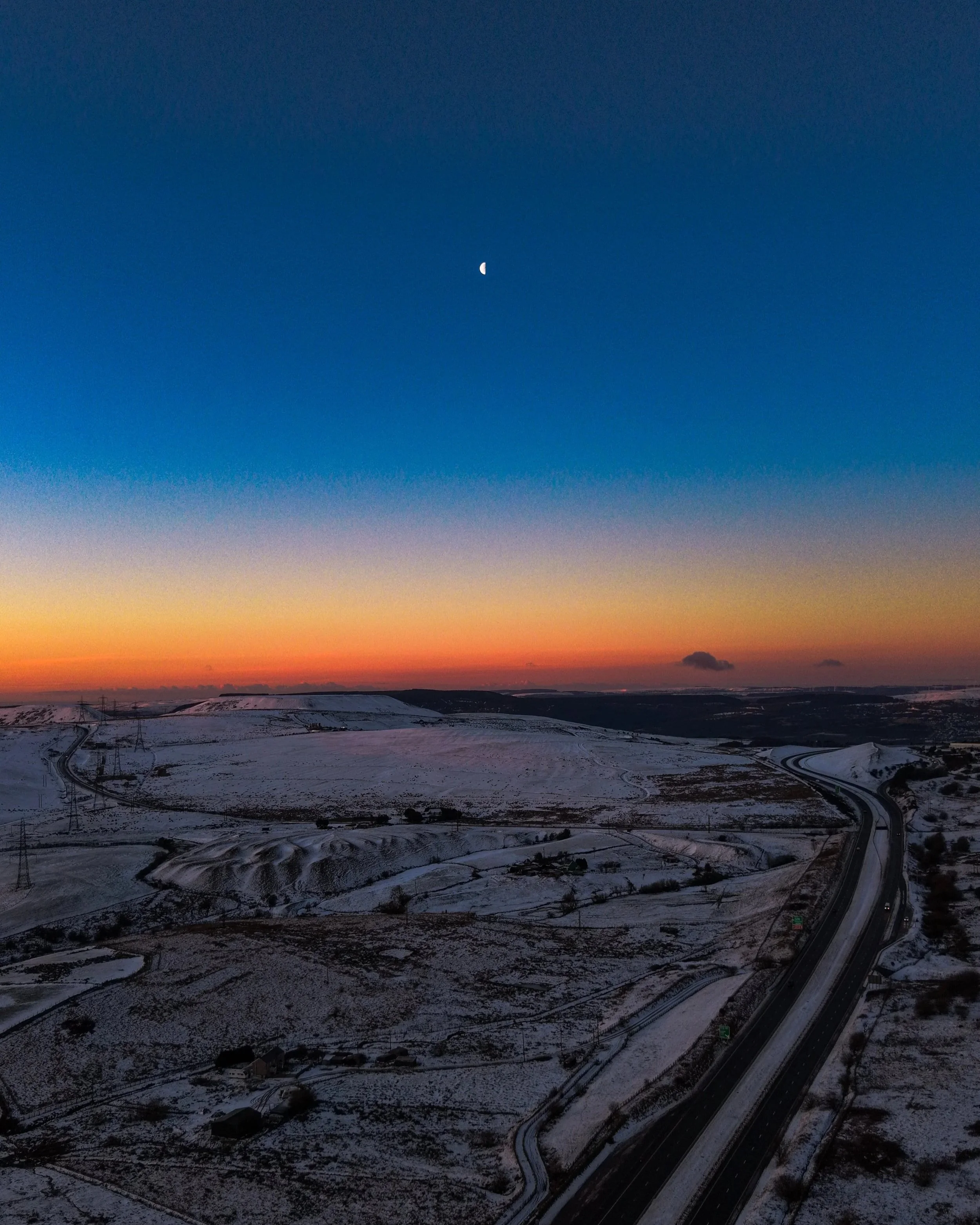 A snow-covered landscape at dusk with a highway on the right side, stretching through hills and fields under a colorful sunset sky with the moon visible.