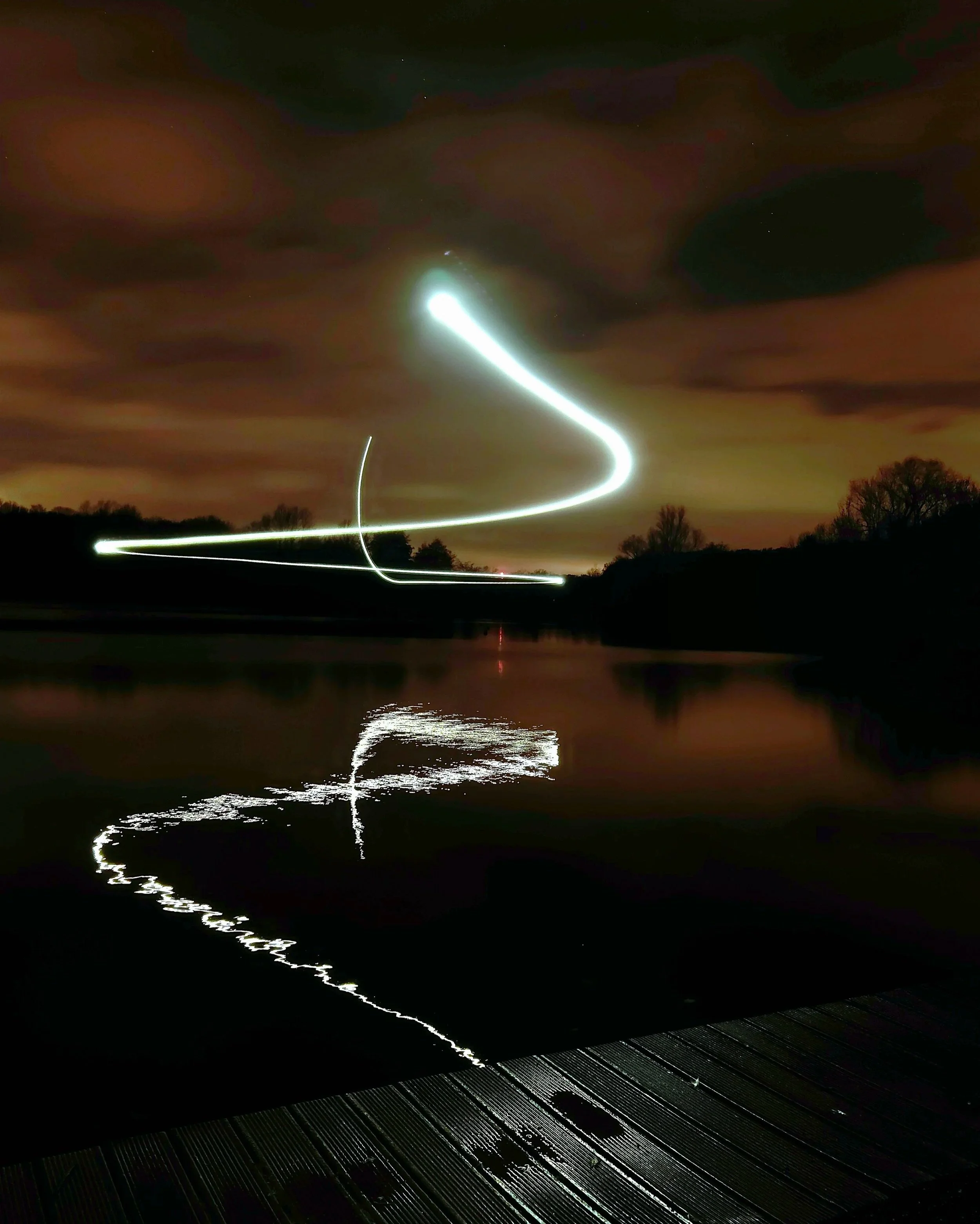 Long exposure photograph of light trails in the night sky over a body of water, with tree silhouettes and a cloudy sky.