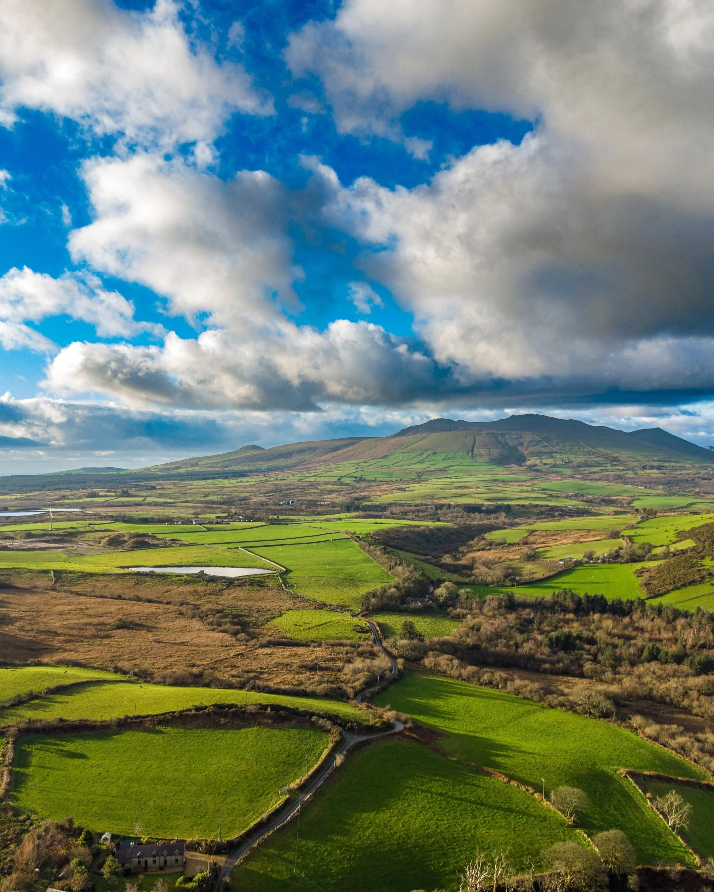 A scenic landscape featuring a large mountain with green slopes, surrounded by patchwork of green fields, small water bodies, and a cloudy blue sky.