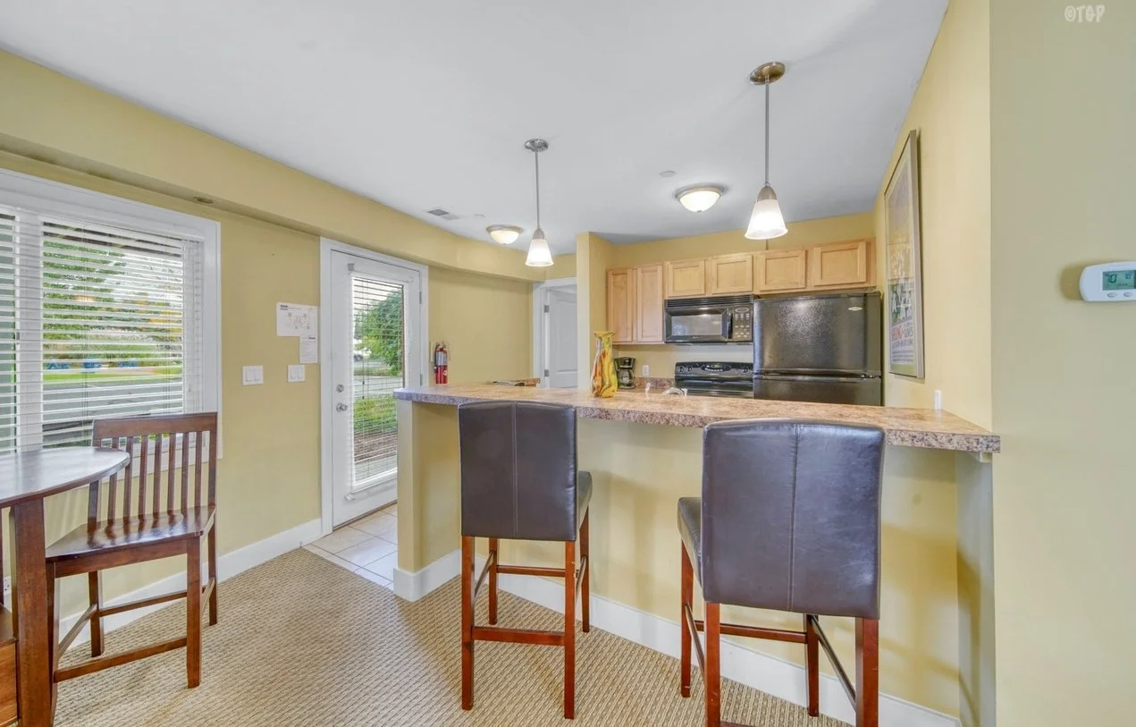 Kitchen with yellow walls, granite countertop breakfast bar, wooden cabinets, black appliances, and seating for two with black chairs. There is a small table and chairs near the window, and a door with a window leads outside.