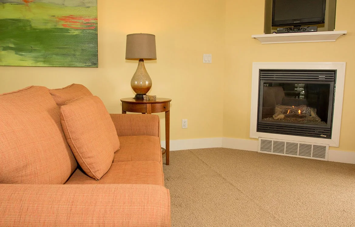 Living room with a peach-colored sofa, a wooden side table with a large lamp, a fireplace, and a wall-mounted TV above the fireplace.