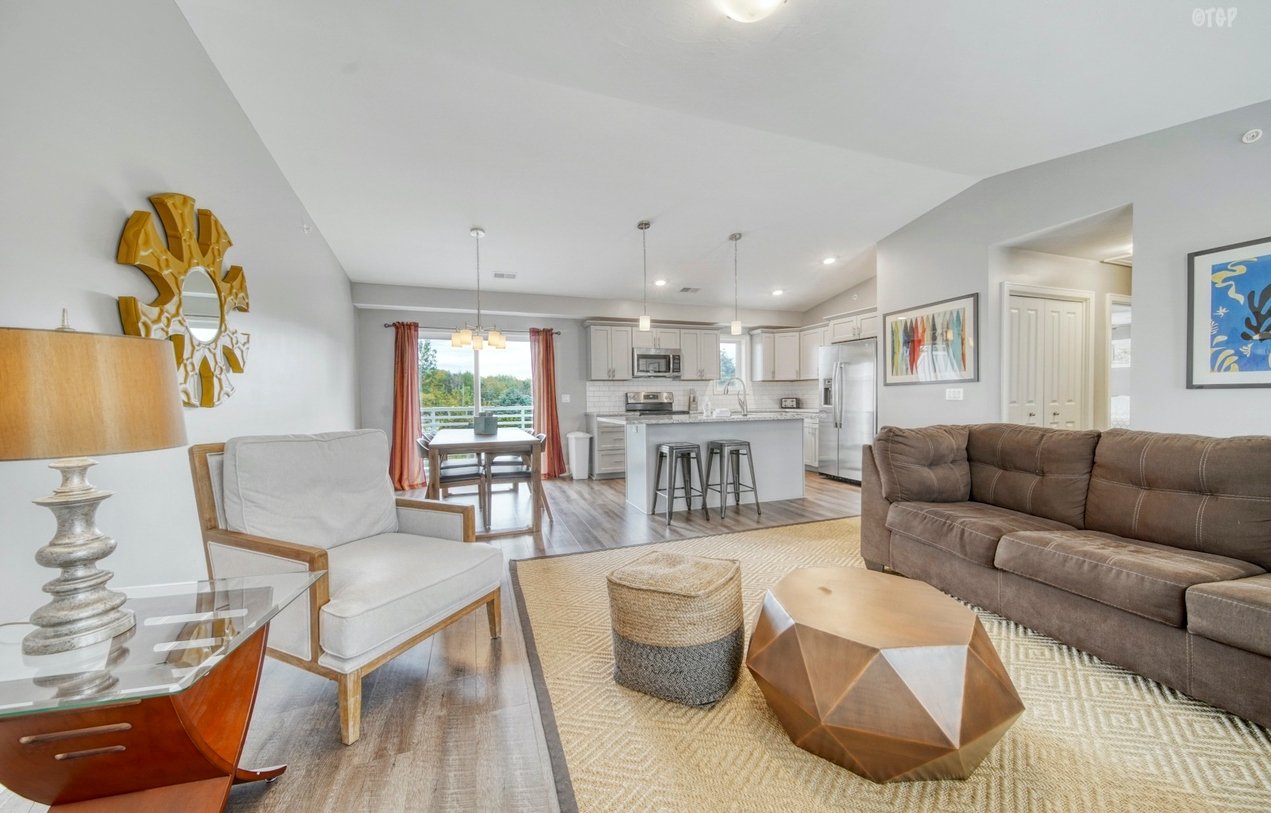 Open-concept living room with brown sofa, white armchair, wooden coffee table, and beige rug, adjacent to a kitchen with white cabinets, island with barstools, and dining area with chandelier and large window with red curtains.