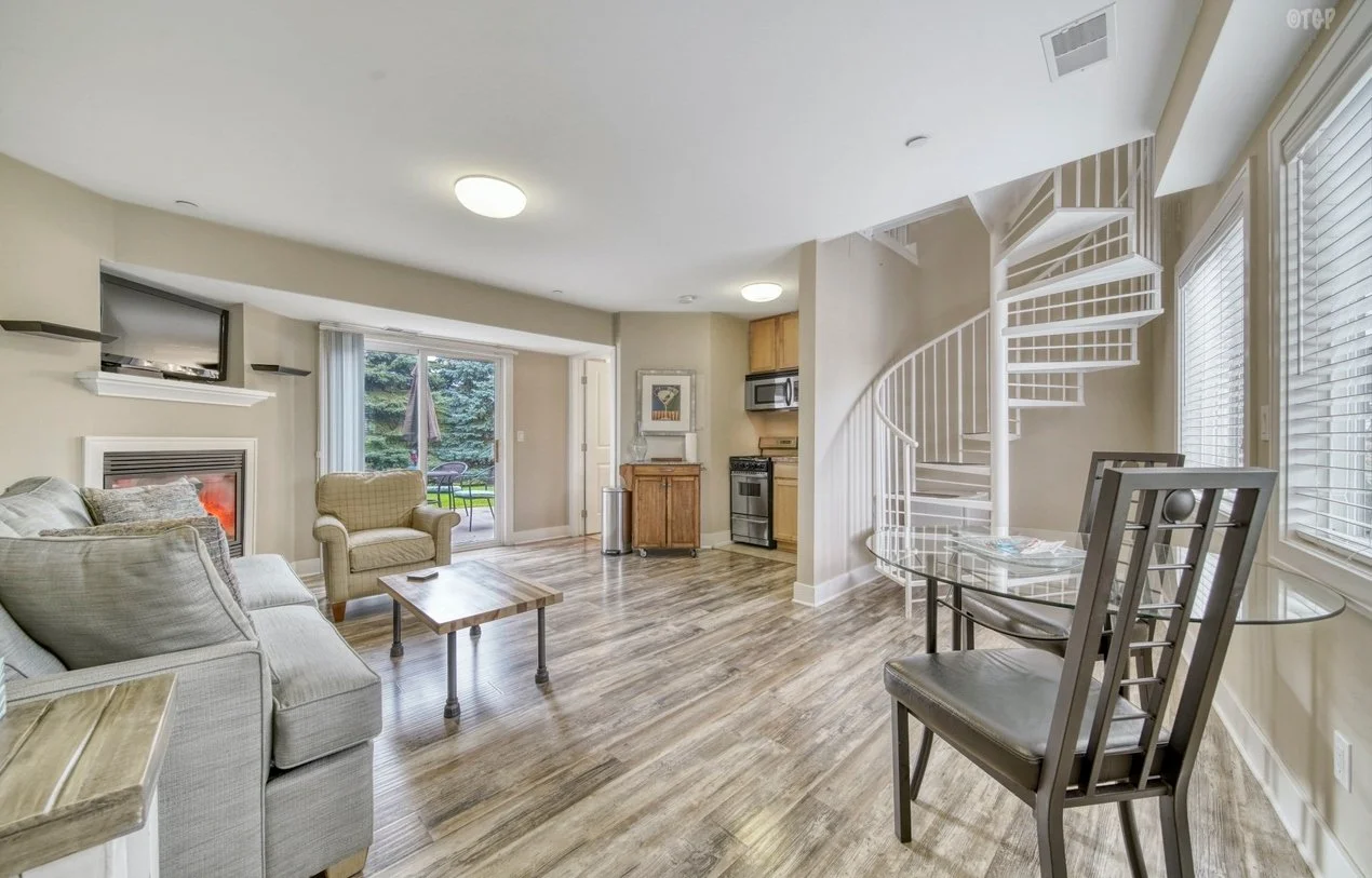 Living room with beige walls, hardwood floors, a gray sofa, a cream armchair, a wooden coffee table, a fireplace, a sliding glass door leading to a patio, a dining table with chairs, a spiral staircase, and large windows with blinds.