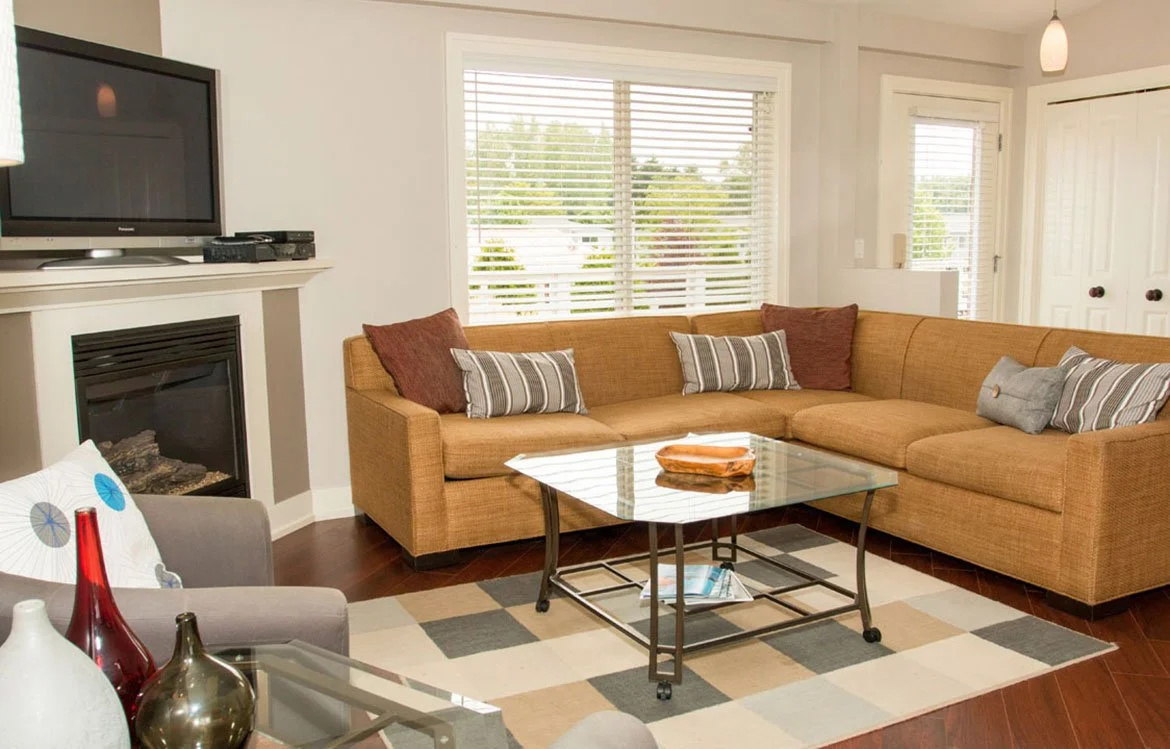 Living room with a large L-shaped brown sofa, striped and solid pillows, a glass coffee table, a fireplace, a flat-screen TV, and large windows with blinds.