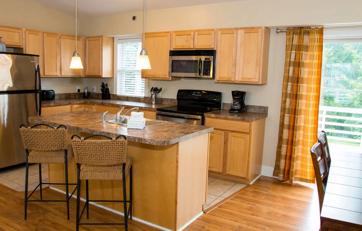 Kitchen with wooden cabinets, granite countertops, stainless steel refrigerator, microwave, oven, coffee maker, and a small kitchen island with two woven bar stools. There is a window above the sink and a sliding glass door with yellow curtains leadi