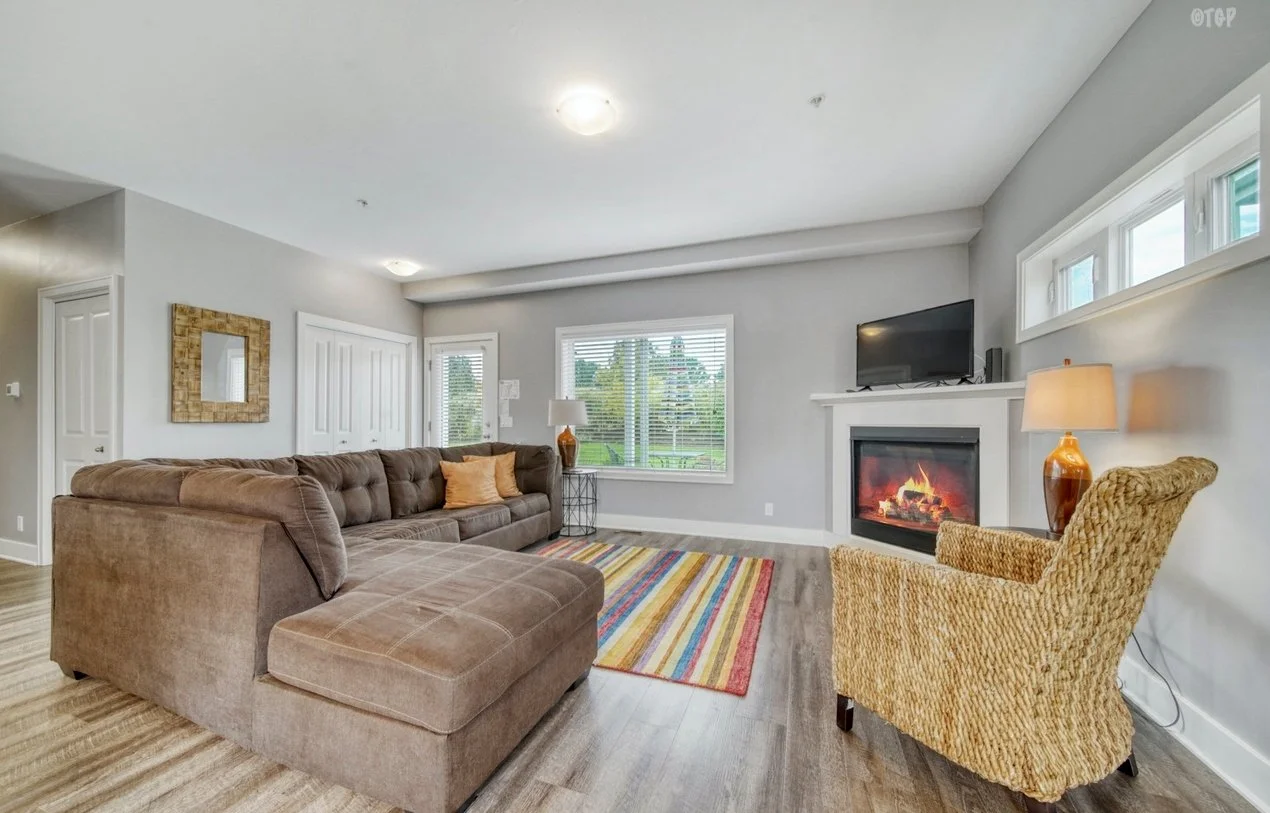 Living room with a brown sectional sofa, a colorful striped rug, a wicker armchair with a lamp, a fireplace with a TV above, and windows showing greenery outside.