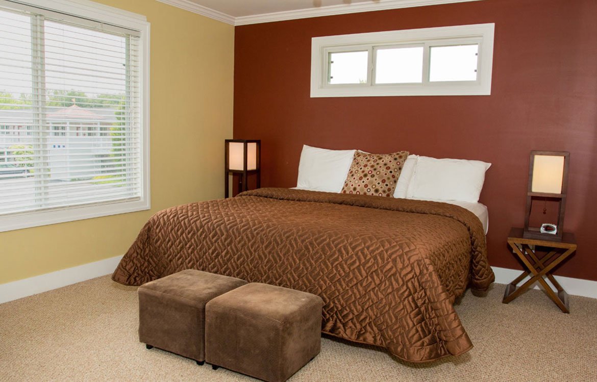 Bedroom with a large bed covered in a brown quilt, two white pillows, one patterned pillow, and two bedside lamps. There are two brown ottomans at the foot of the bed, a window with blinds on the left, and a small rectangular window above the bed on 