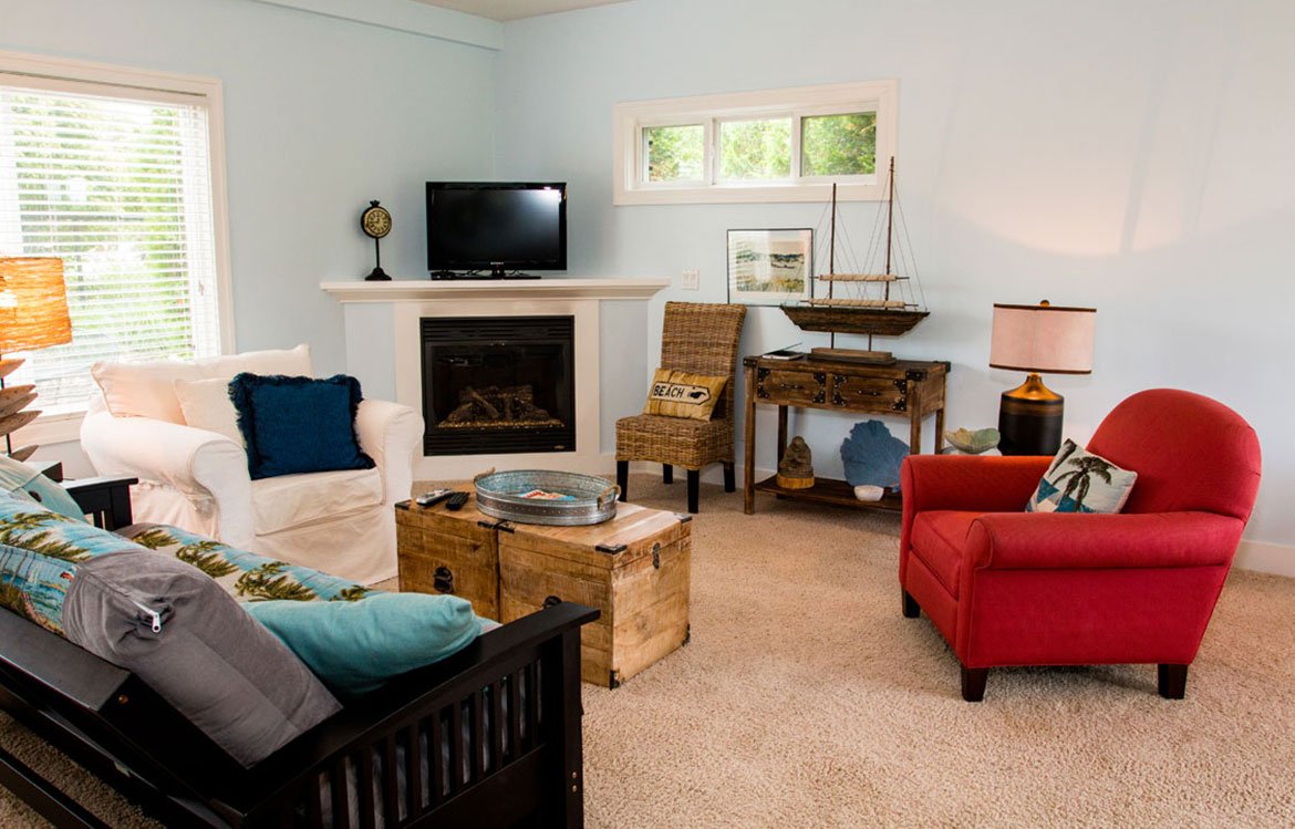 Cozy living room with white and red armchairs, a wooden coffee table, a fireplace with a TV above, a wicker chair, and beach-themed decor, including a maritime ship model and framed artwork. Bright natural light filters through the windows.