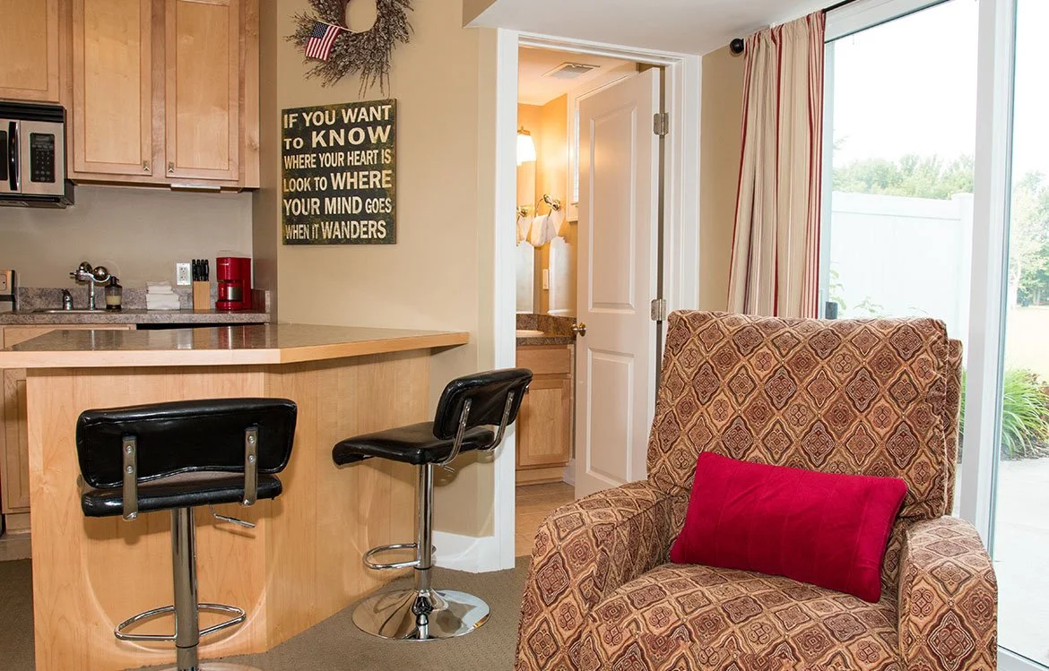 Interior view of a cozy living area with a patterned armchair with a red pillow, a small kitchenette with wooden cabinets, a breakfast bar with two black stools, a door leading to a bathroom, and a large sliding glass door with curtains opening to a 