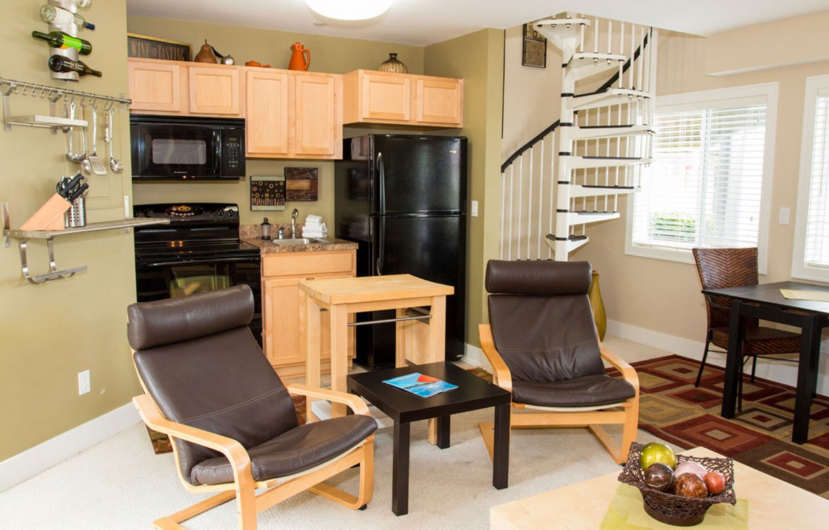 Living room and kitchen area with black appliances, wooden cabinets, spiral staircase, armchairs, window, and dining table.