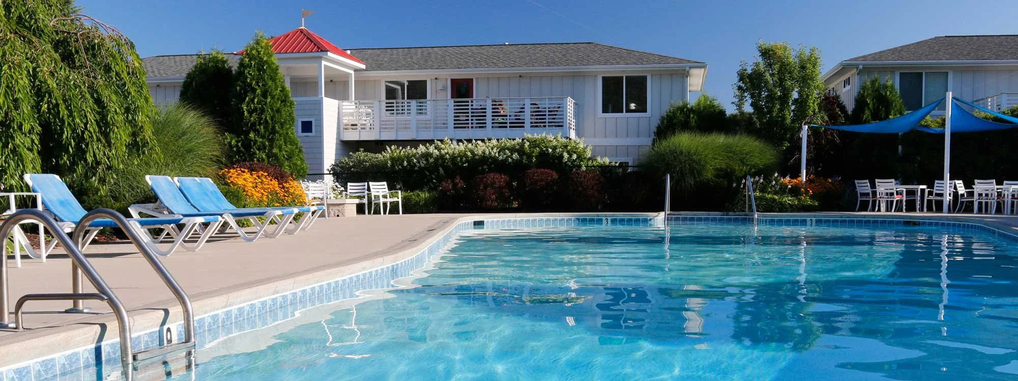 Swimming pool with blue lounge chairs and white tables beside bushes and trees, apartment building with a balcony and red roof in the background, shaded seating area with blue canopy.