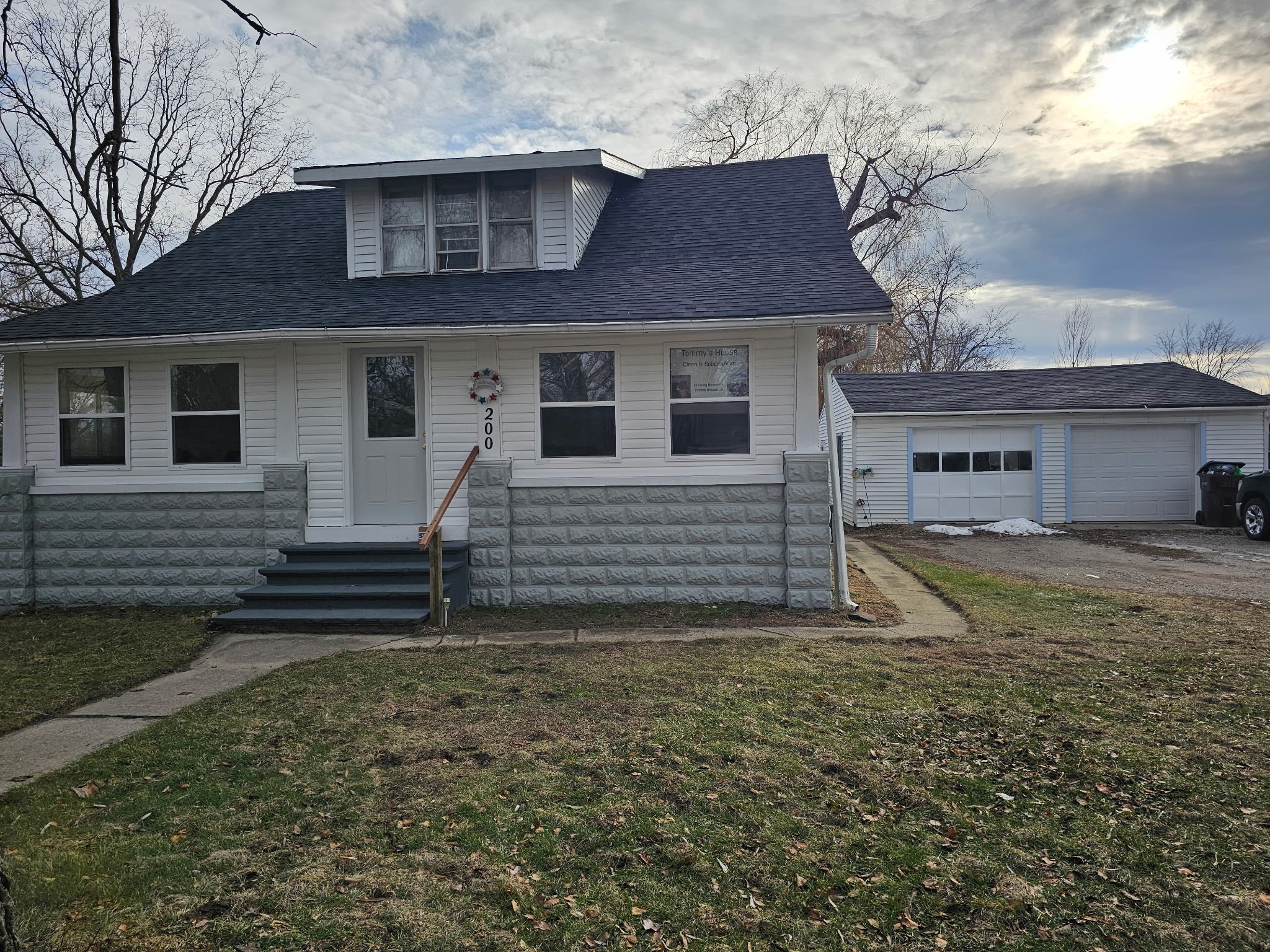 Front view of a white, two-story house with a grey stone foundation, black stairs, and a black roof, surrounded by leafless trees and a grassy lawn.
