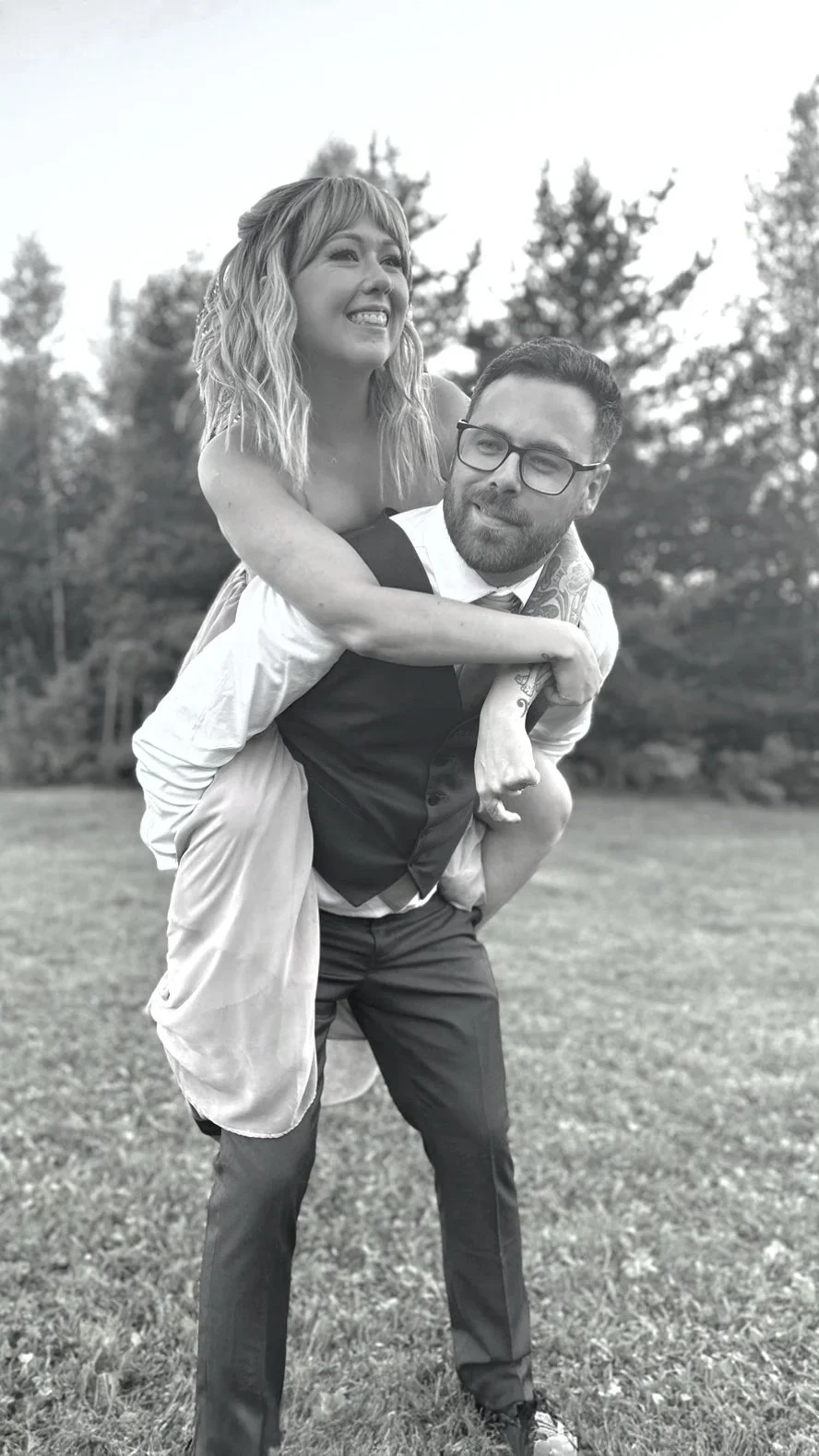 A black and white photo of a woman with wavy hair on someone's back, outdoors on a grassy area, smiling, with trees in the background.