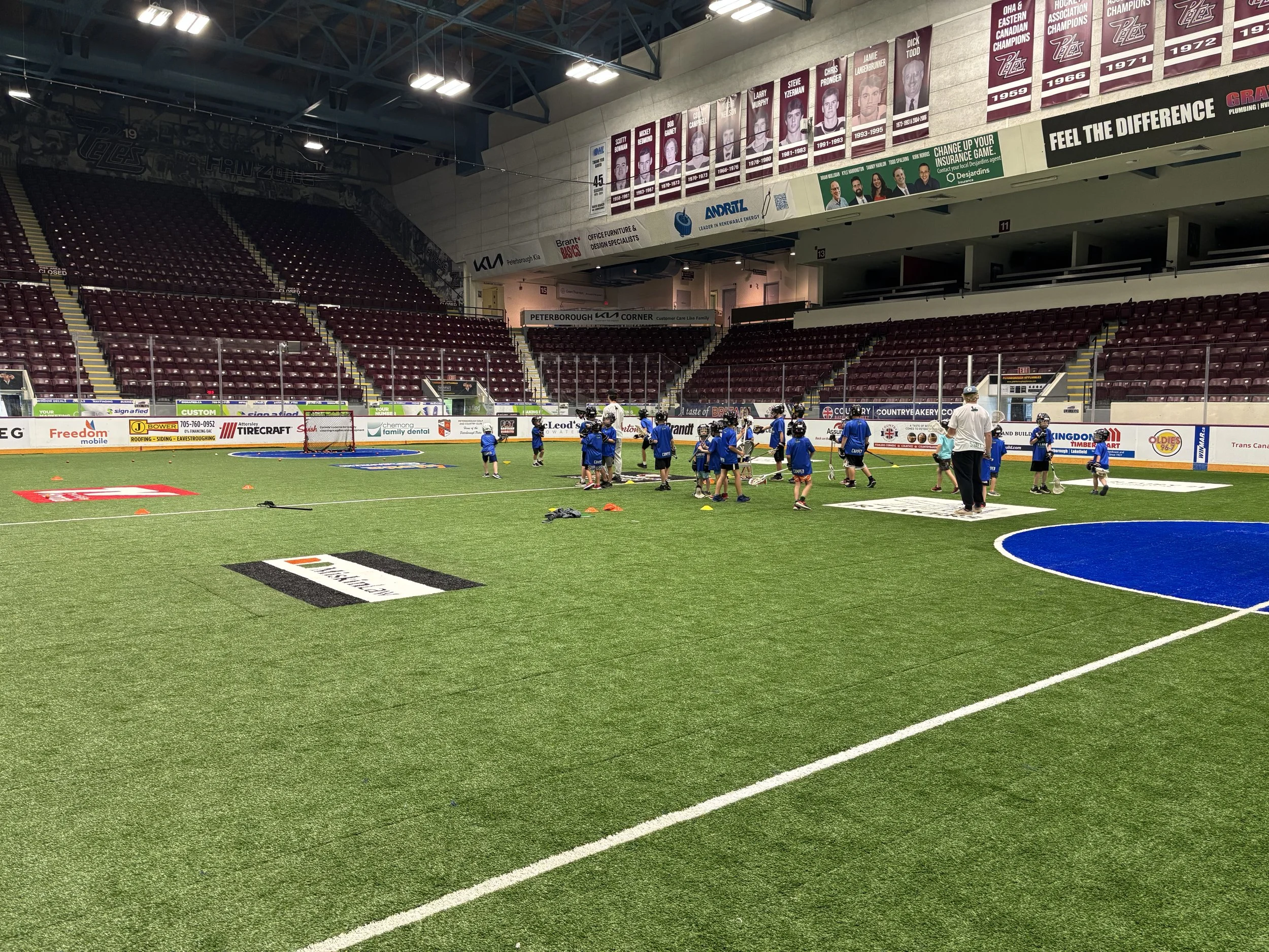 Indoor arena with a turf field where young kids in blue sports uniforms are practicing lacrosse, supervised by coaches, with banners and empty red seats in the background.
