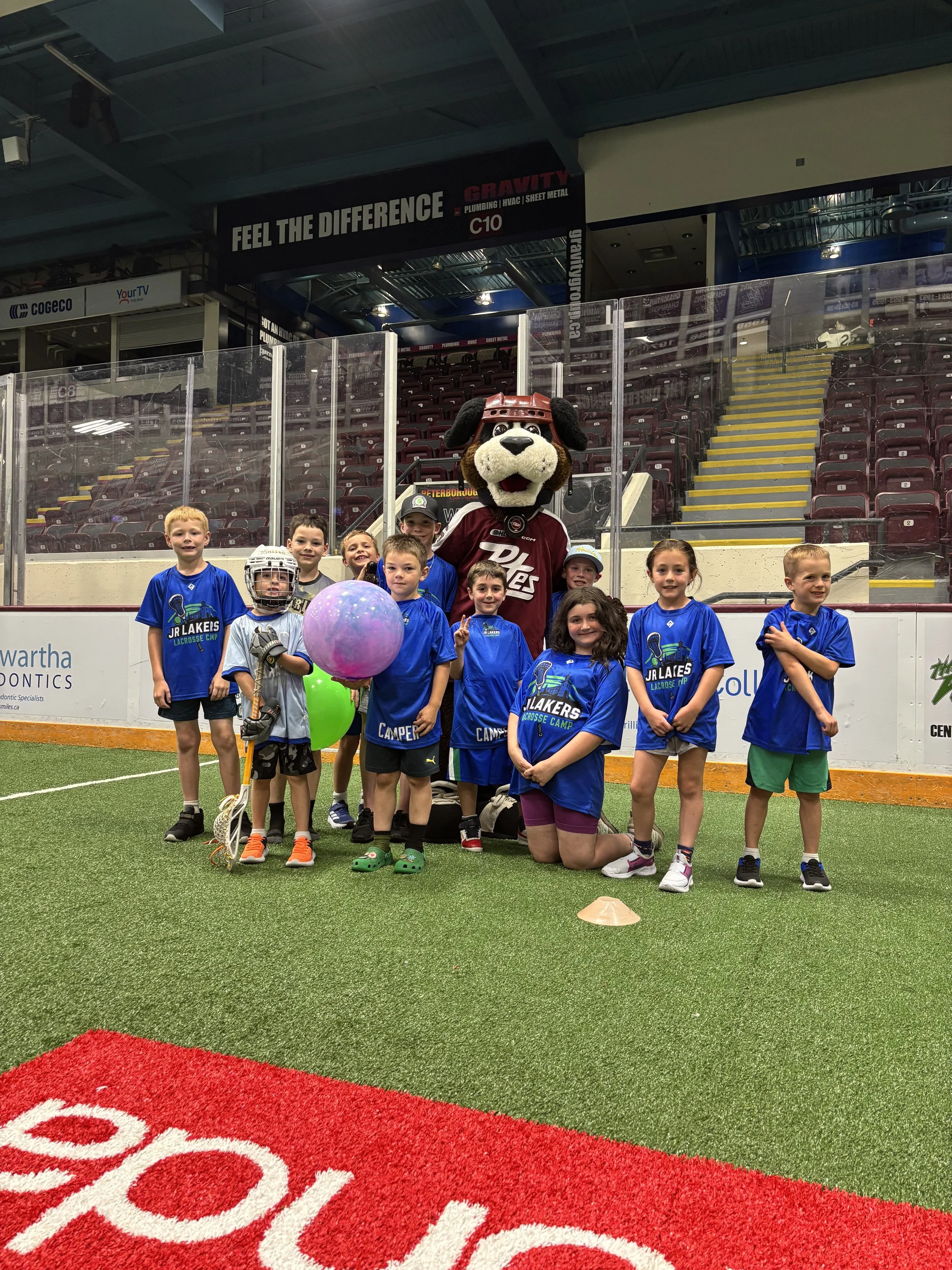 Children and a mascot posing on an indoor sports field, some holding balloons, with a hockey stick, and dressed in sports uniforms.