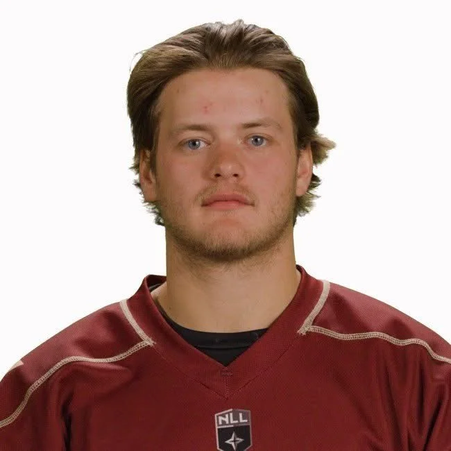 Young man with light skin, medium-length brown hair, and facial hair wearing a maroon hockey jersey with the NLL logo.