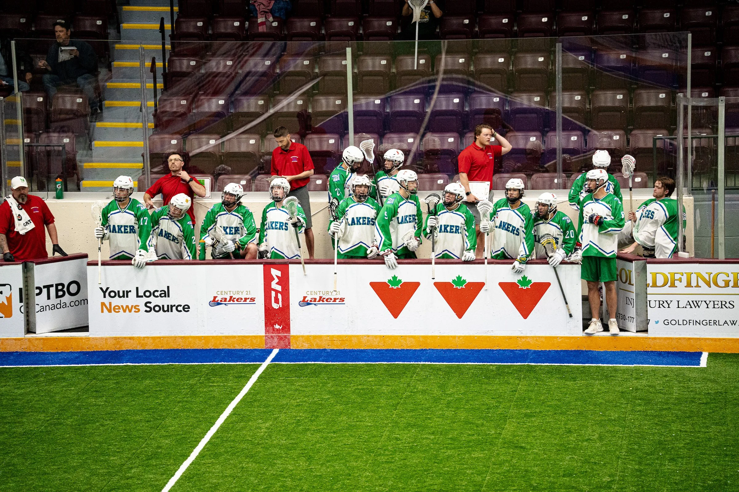 Lacrosse team in green and white uniforms on the bench during a game, with coaches and spectators visible in the background.