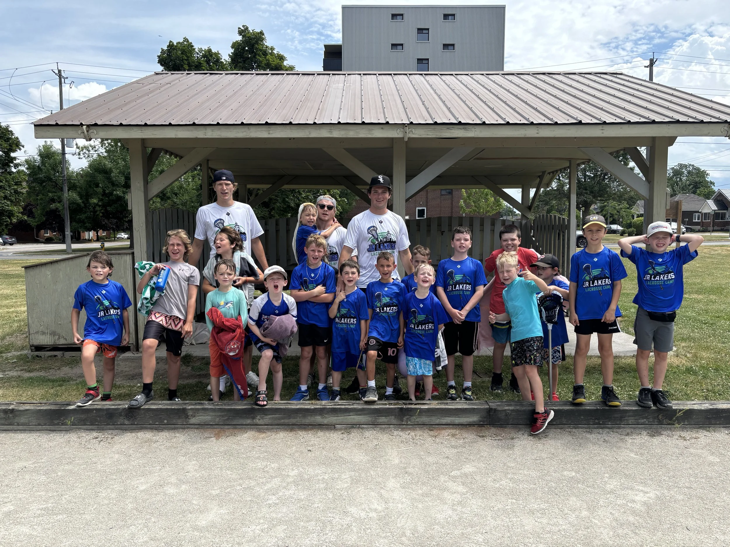 A group of children and adults posing for a photo under a pavilion outdoors, many children wearing blue T-shirts with a lacrosse logo, on a sunny day with a grassy field and buildings in the background.