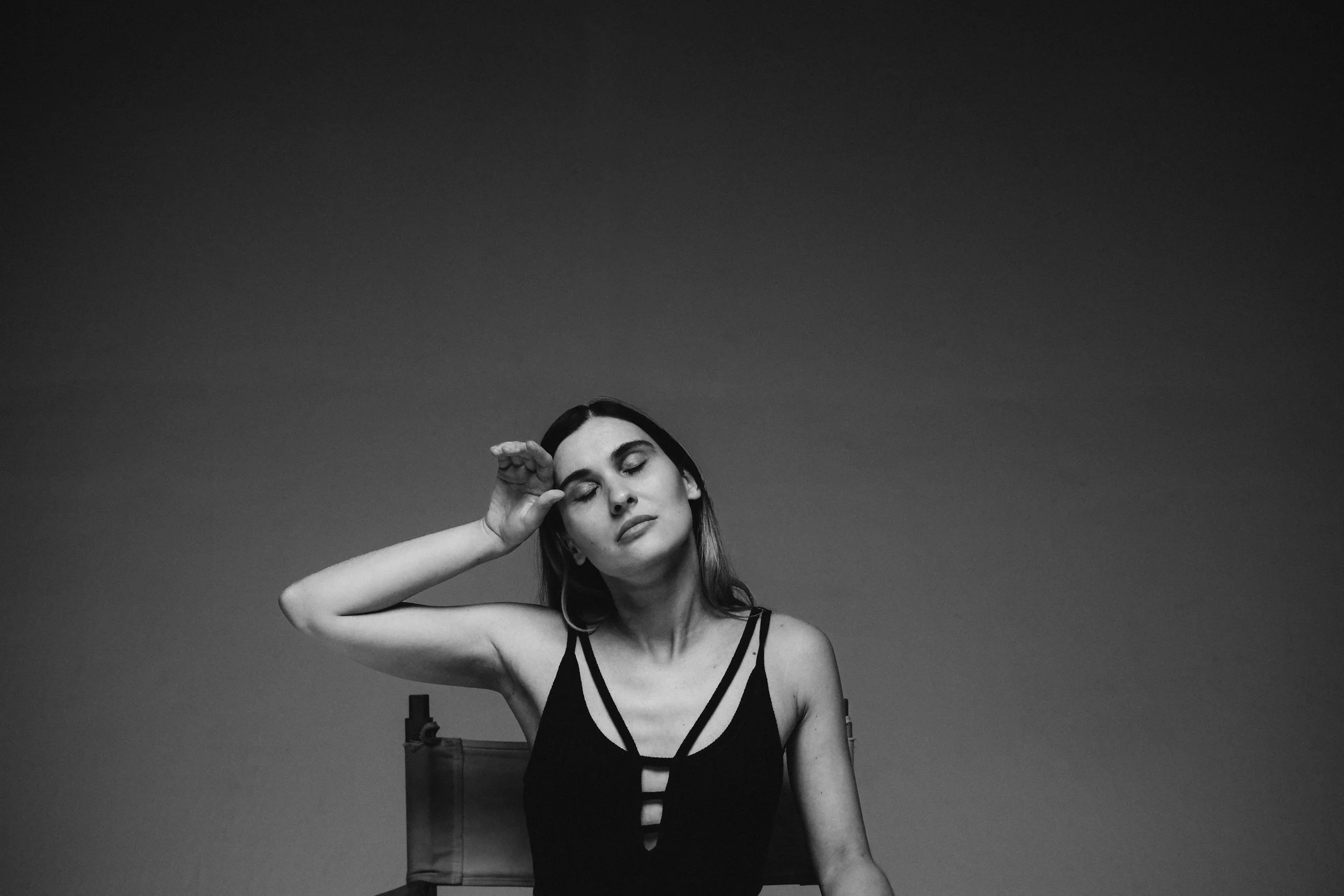 Mujer de cabello largo, vestida con un vestido negro con recortes, con los ojos cerrados y una mano en la frente, en un fondo sin detalles, en una fotografía en blanco y negro.
