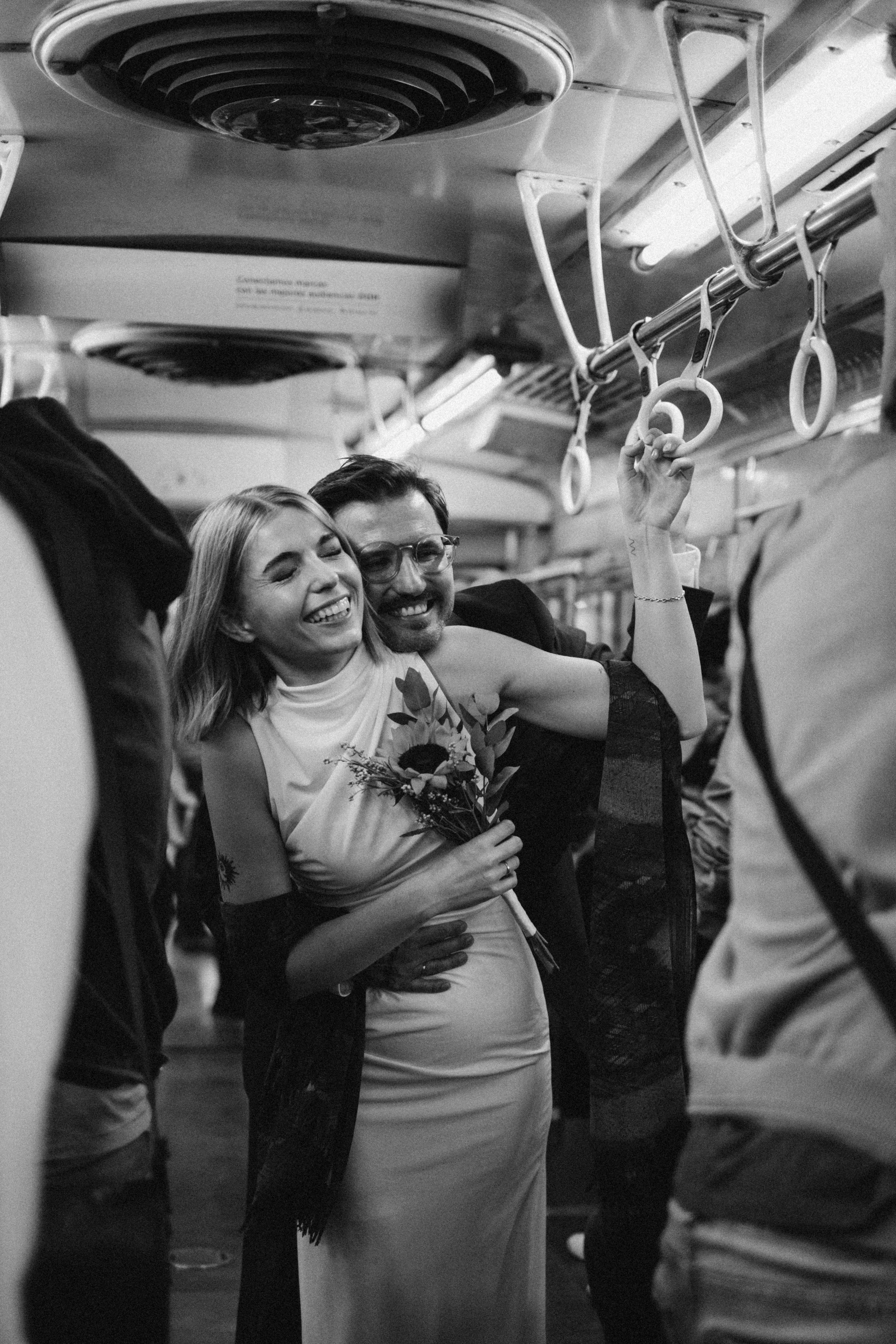 Pareja abrazada y sonriendo en un transporte público, ella sostiene un ramo de flores y ropa formal, él lleva gafas y ropa casual, mientras otros pasajeros los rodean y hay barras para agarrarse.
