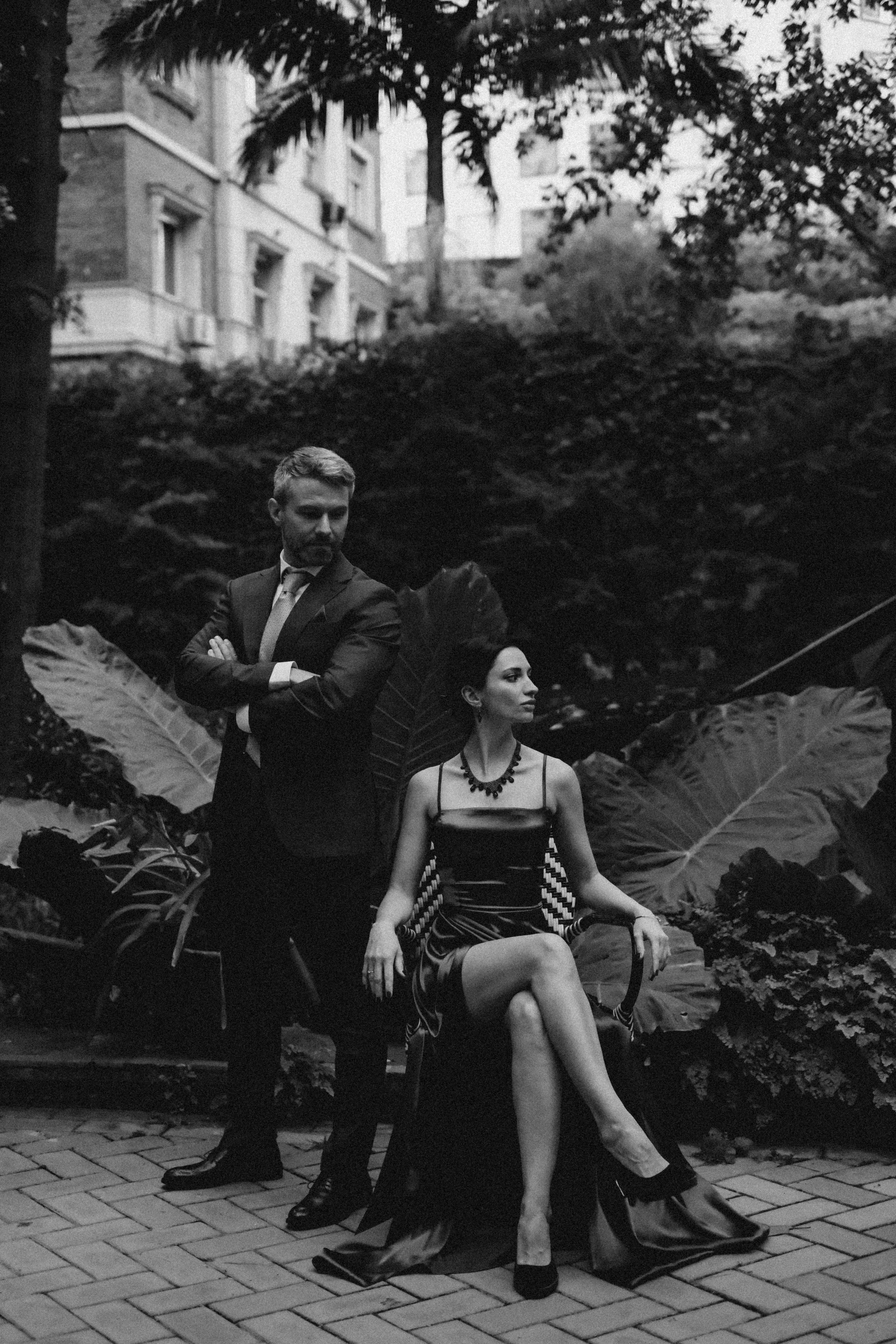 Una mujer sentada en una silla con vestido largo, acompañada de un hombre de traje, en un entorno natural con plantas grandes y árboles, en un espacio que parece ser un parque o jardín.