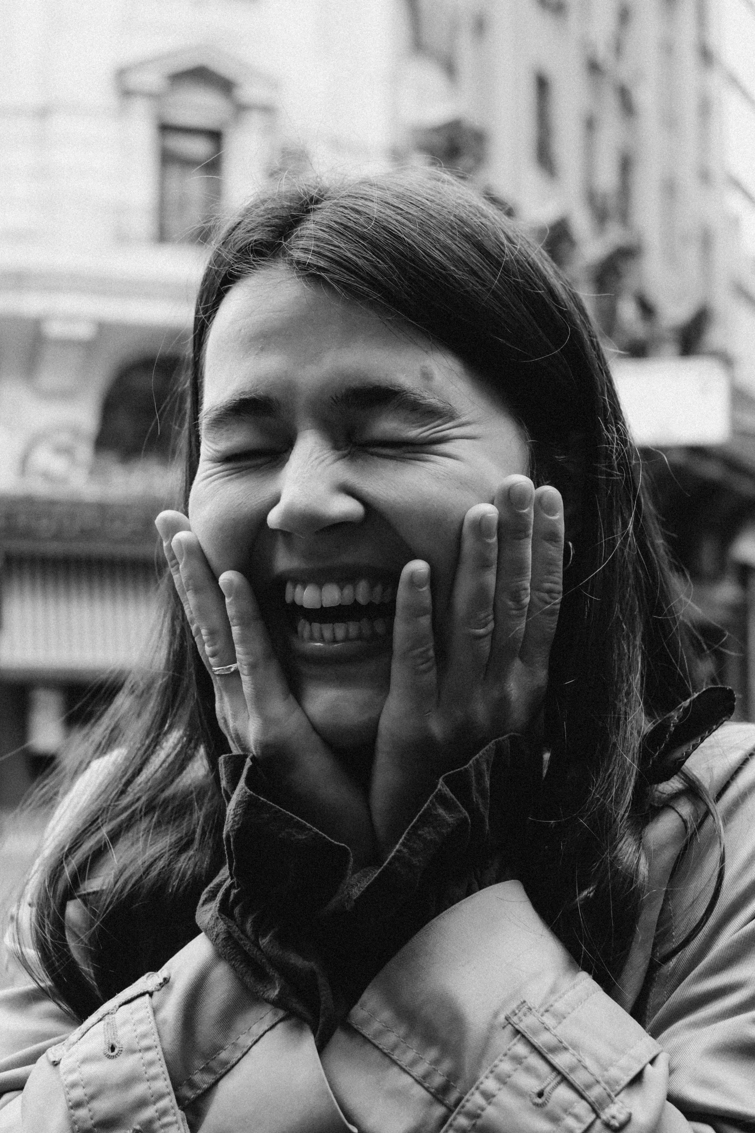 Mujer sonriendo con manos en las mejillas, en un entorno urbano, en blanco y negro.