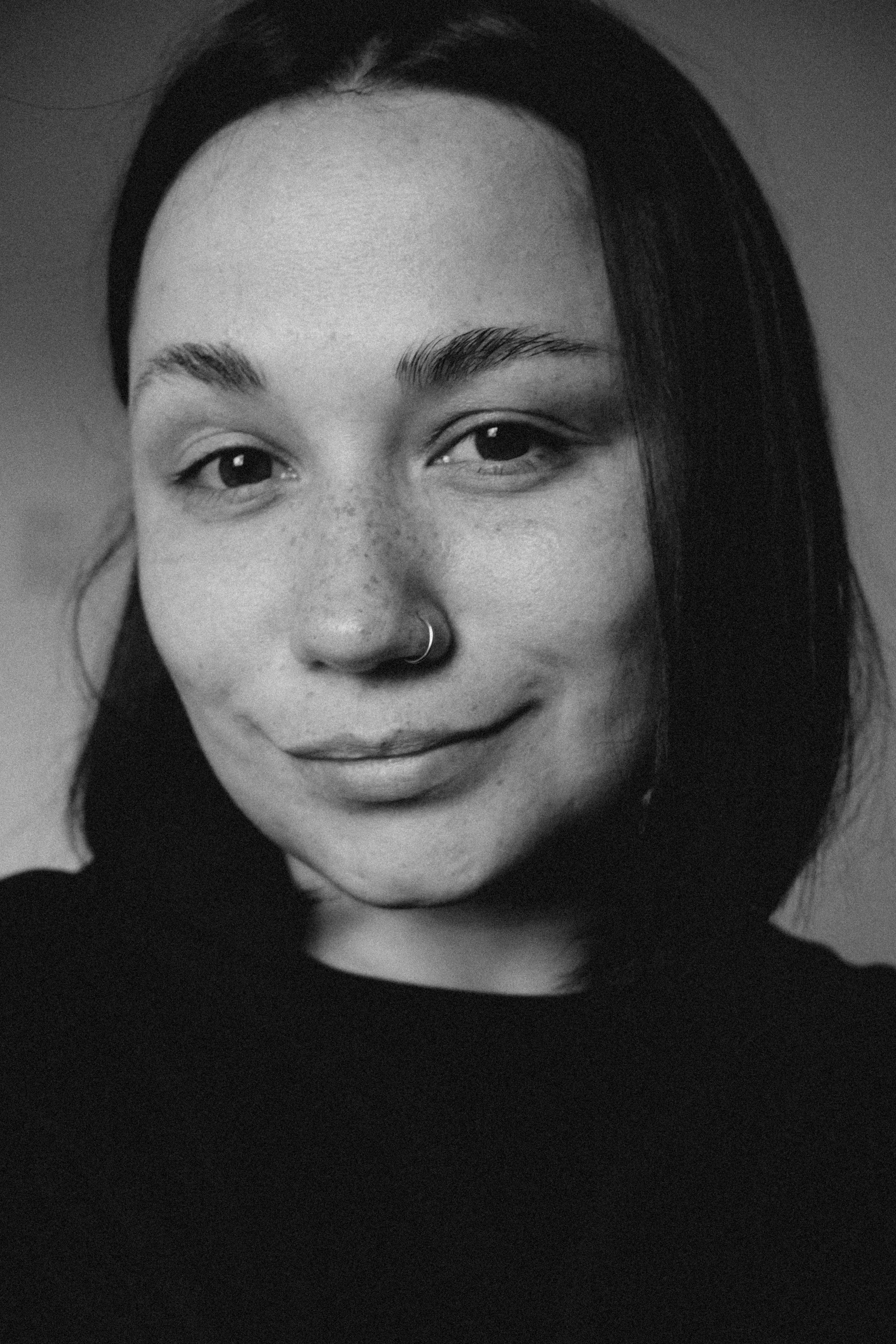 A black and white close-up portrait of a woman with straight, shoulder-length hair, a nose ring, and a subtle smile.