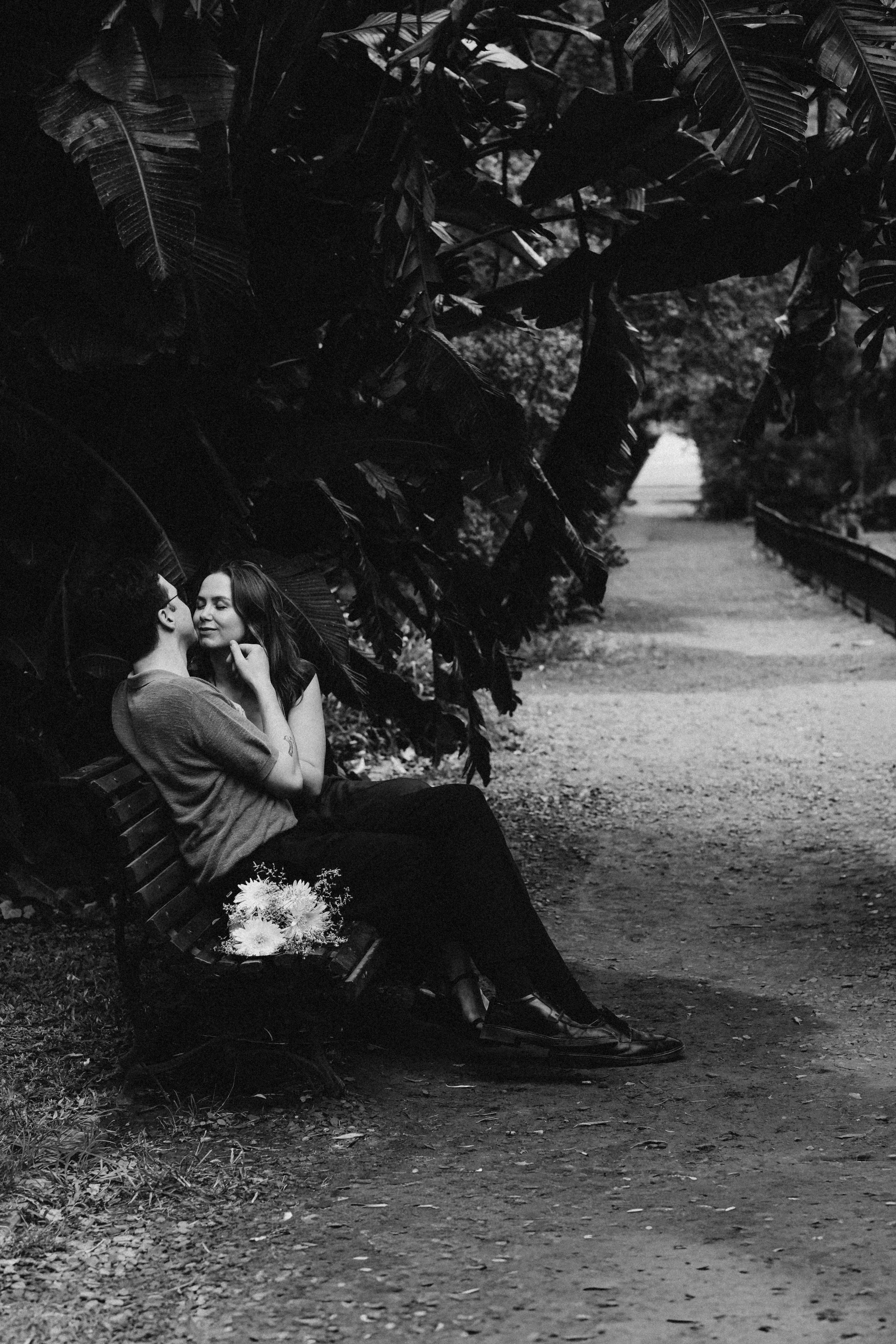 Una pareja sentada en un banco en un parque, ella le toca el rostro y se miran con cariño, entre las hojas grandes y oscuras de los árboles que rodean el sendero.