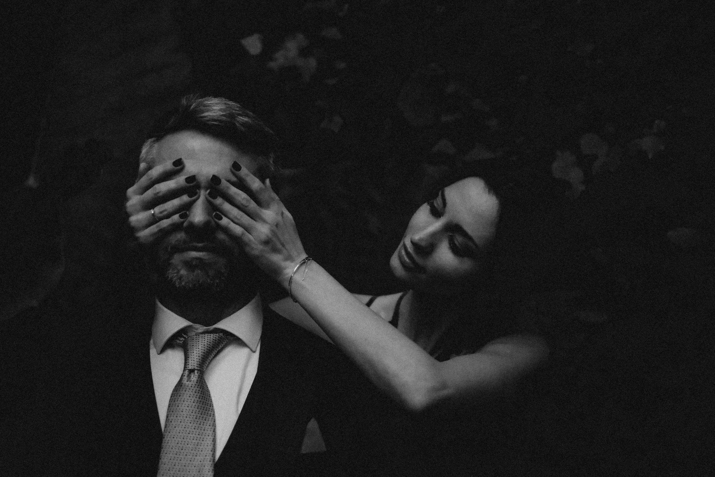 Una pareja de adultos está en un ambiente oscuro y la mujer cubre los ojos del hombre con sus manos.