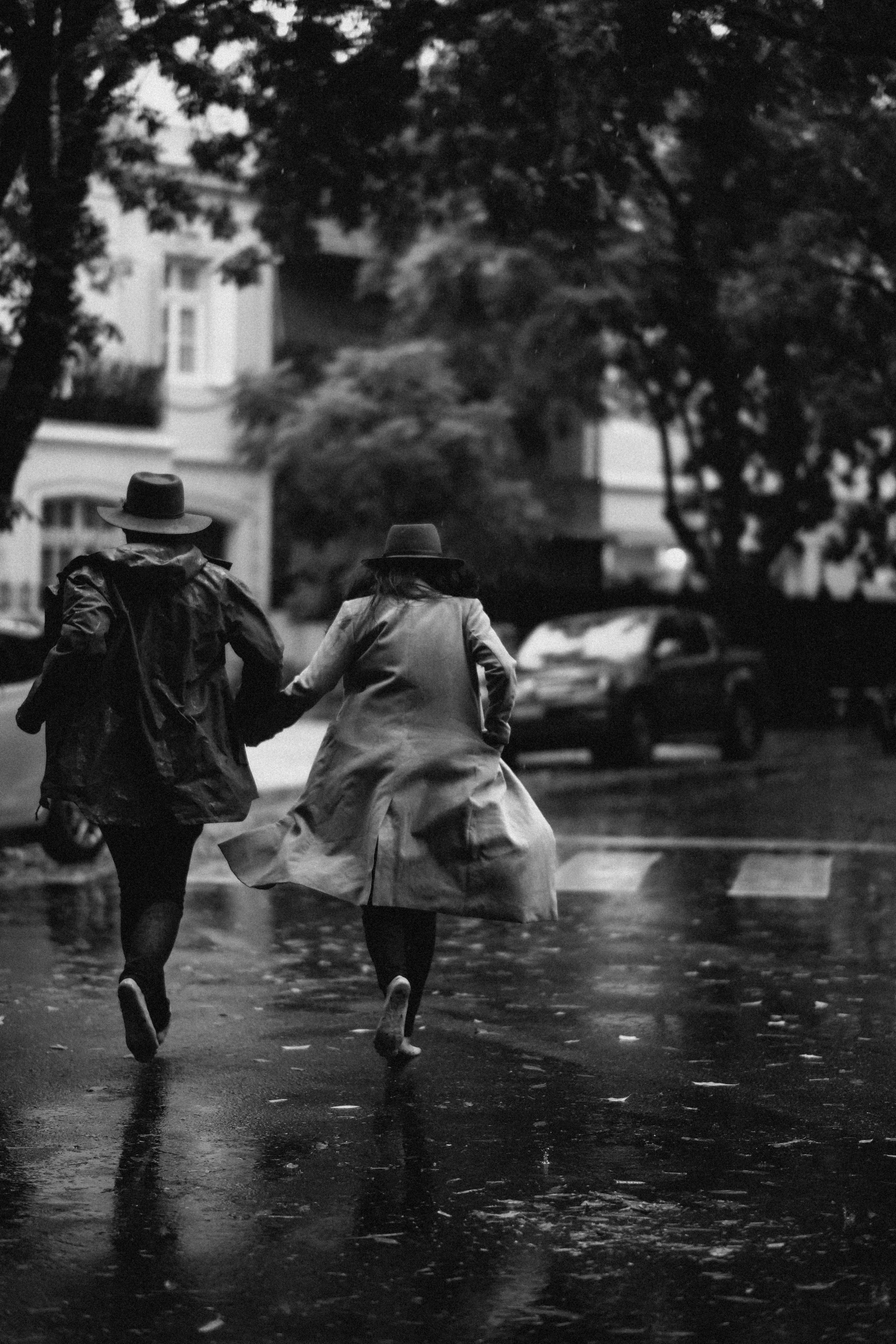 Dos personas caminando por una calle lluviosa, sosteniéndose de la mano, con árboles y casas al fondo, en un día sombreado.