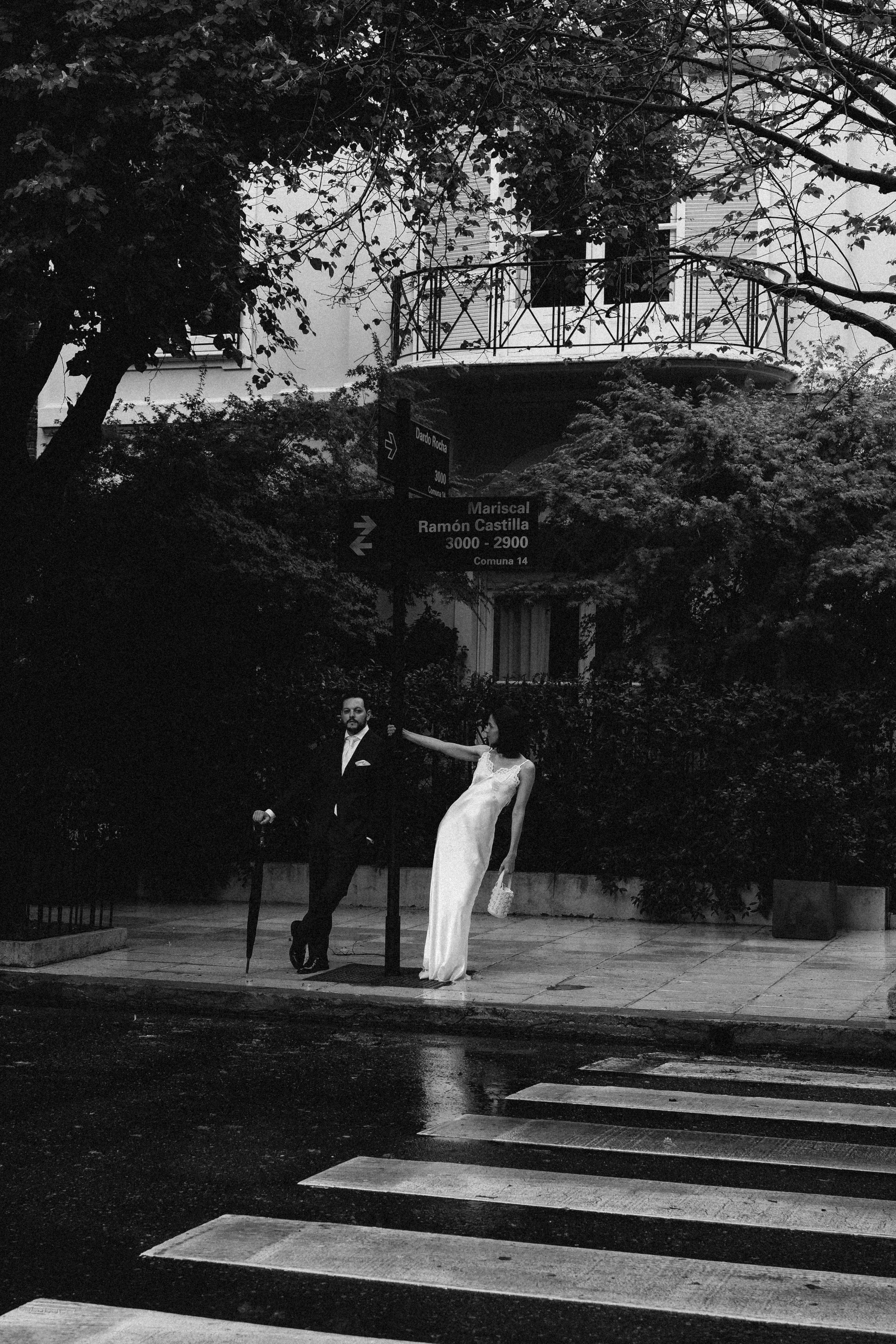 Pareja vestida de boda caminando por la calle, ella lleva vestido largo blanco y él un traje negro, con un paraguas en la mano, en un entorno urbano con árboles y edificios.