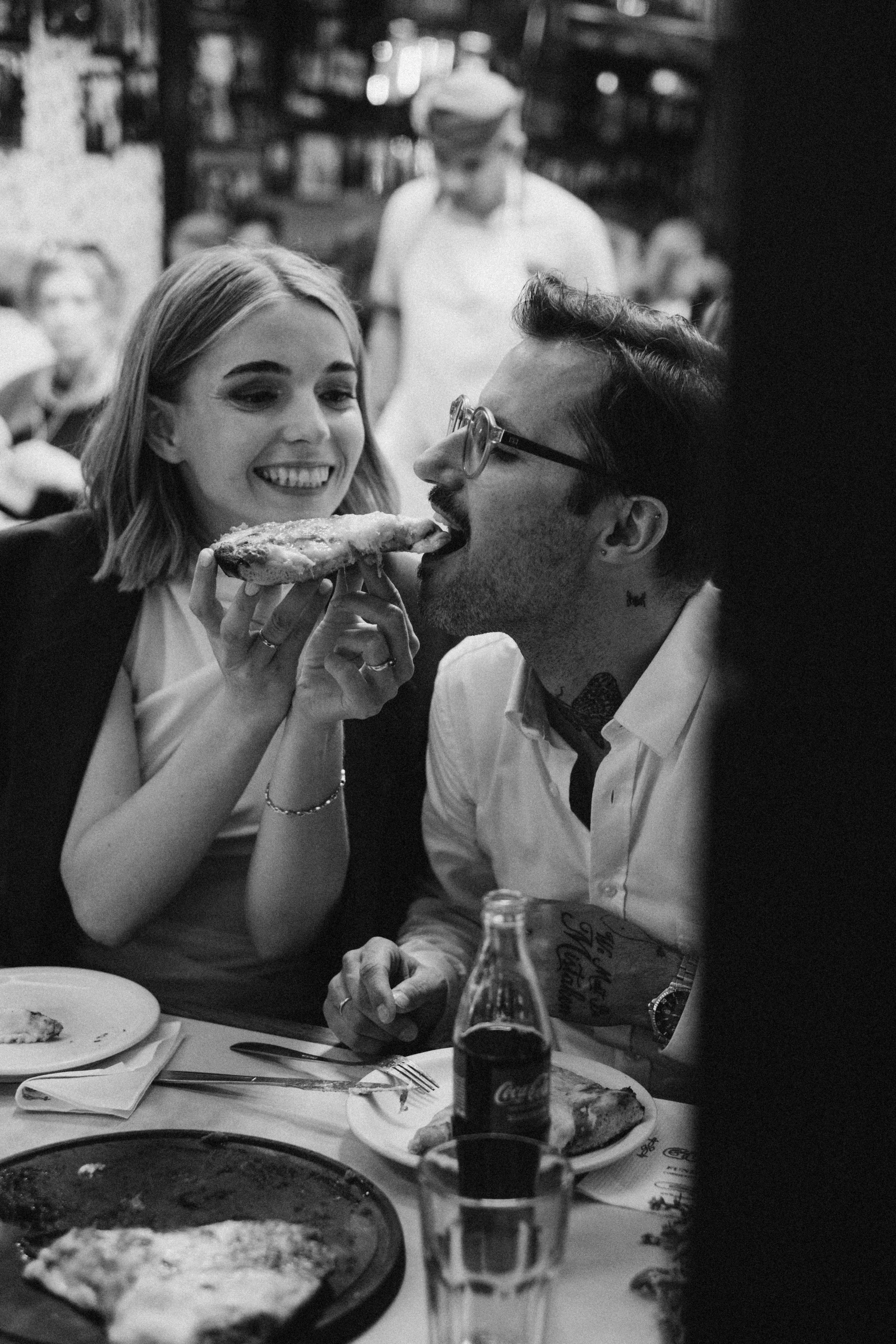 Una pareja en un restaurante compartiendo una rebanada de pizza. Hay una botella de Coca-Cola en la mesa. Fondo con otras personas y estantes con libros o objetos decorativos.