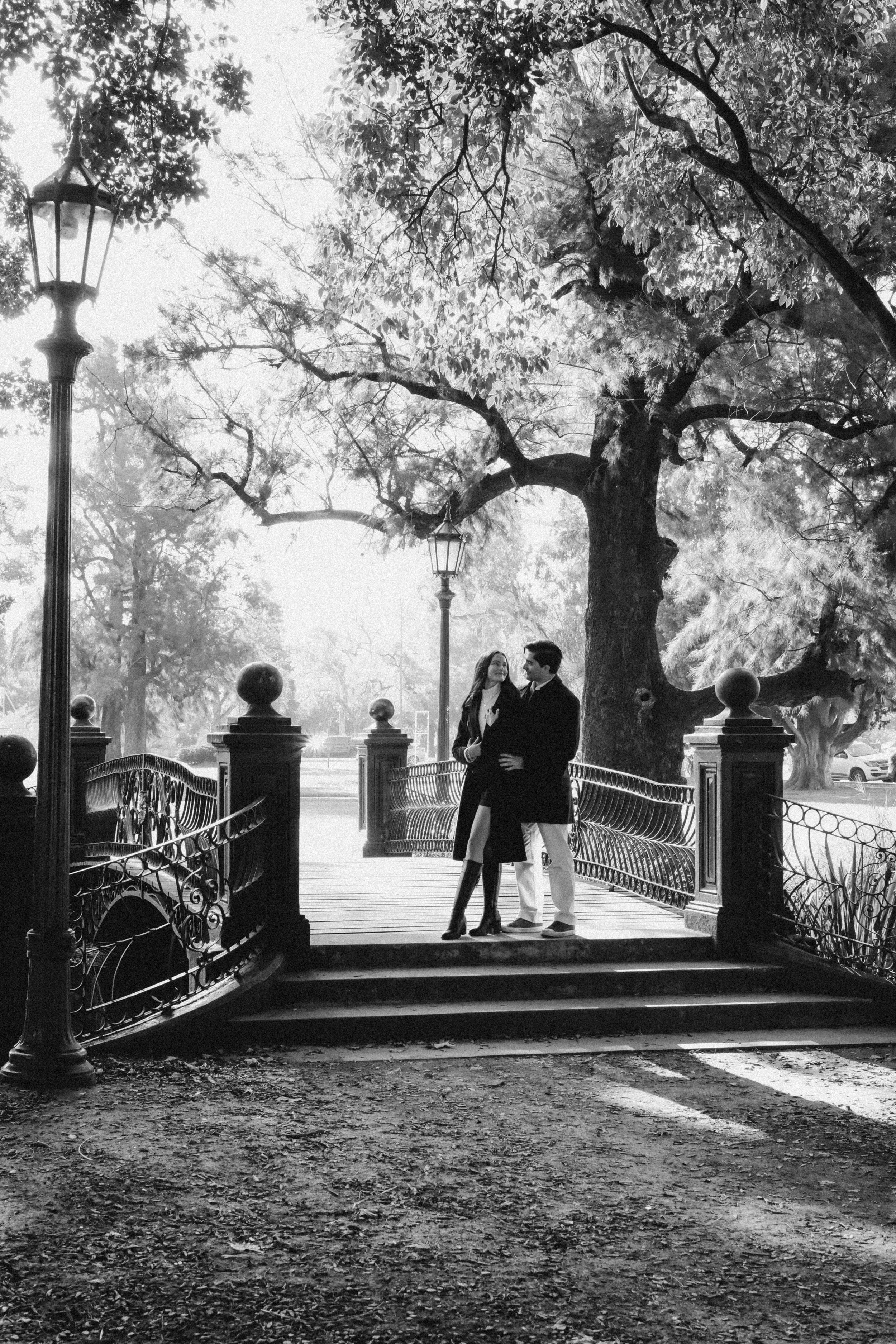 Una pareja de jóvenes en la entrada de un parque con árboles y faroles, en una escena en blanco y negro.