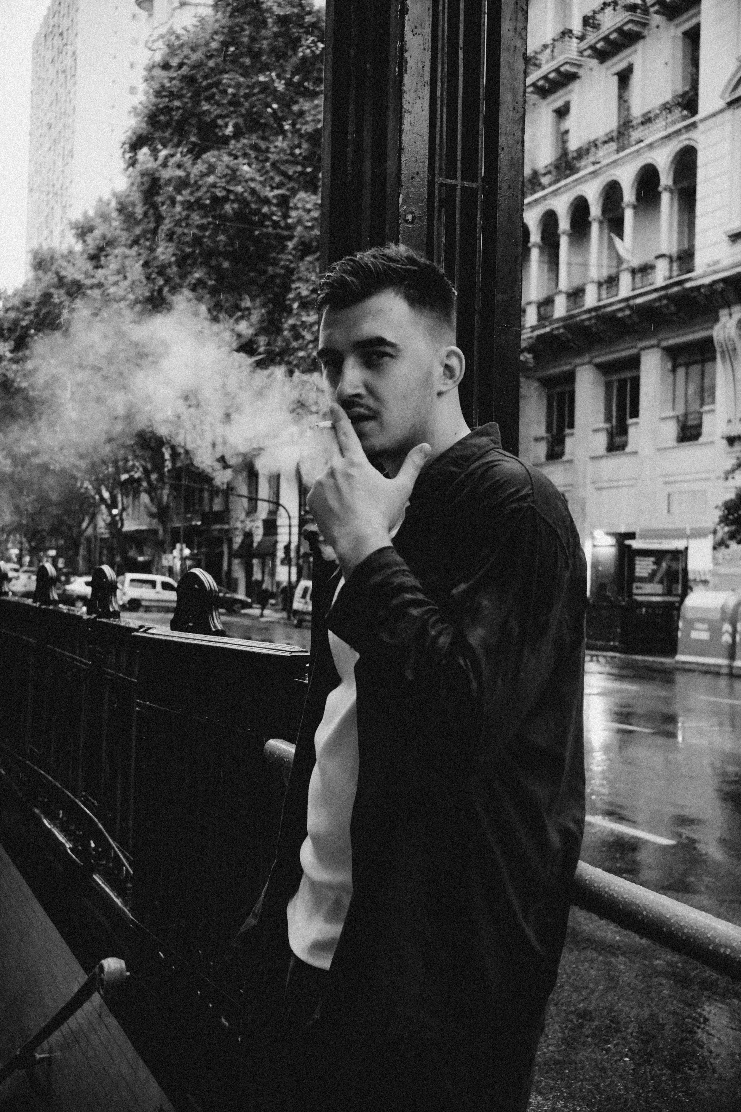 Joven hombre con cabello corto y barba, sosteniendo un cigarrillo y un encendedor, de pie junto a una reja en una calle urbana con edificios y árboles en el fondo, en una foto en blanco y negro.