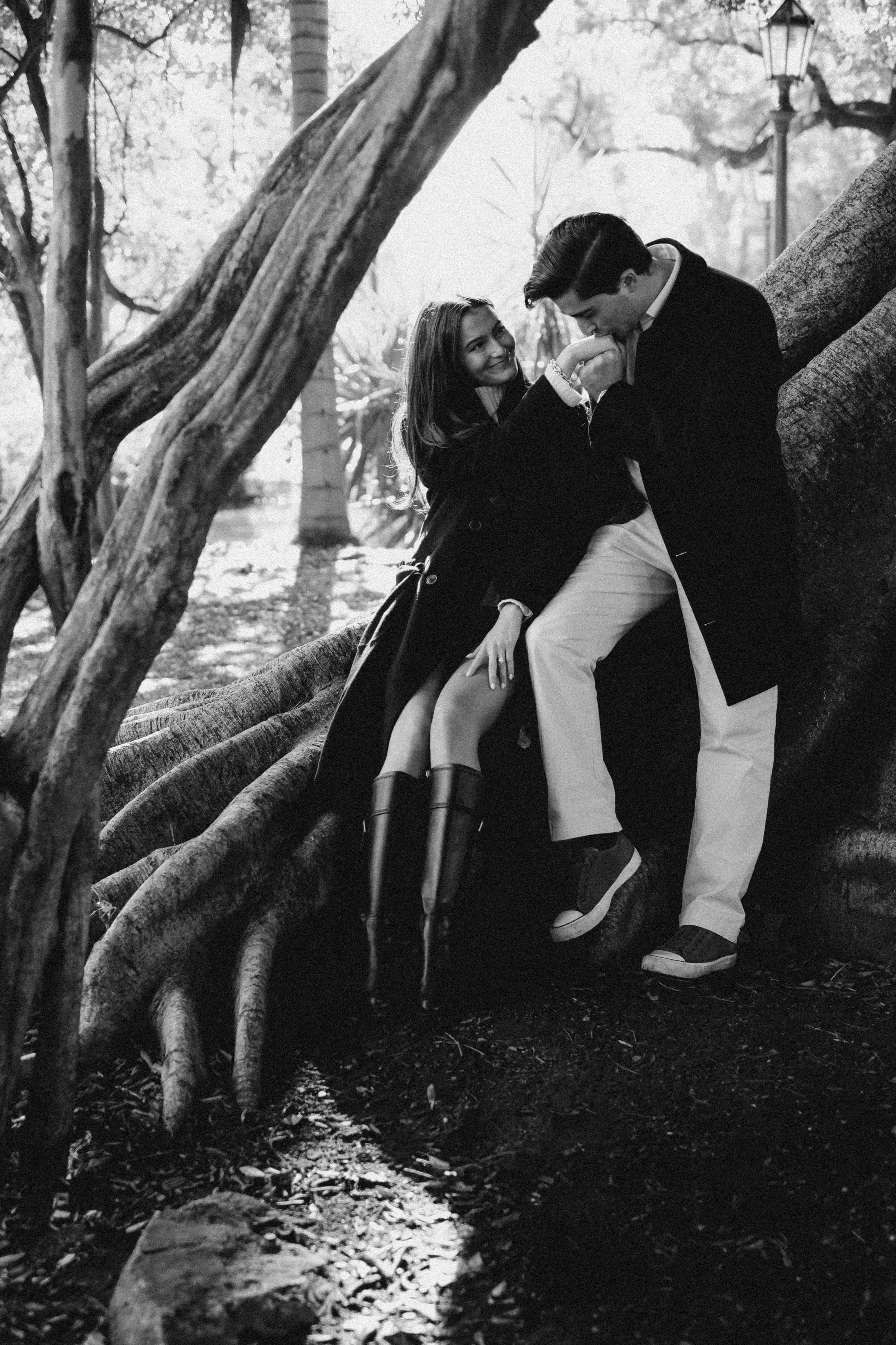 Una pareja en un parque, el hombre está inclinándose para besar la mano de la mujer, ella sonríe, ambos usando ropa elegante y rodeados de árboles.