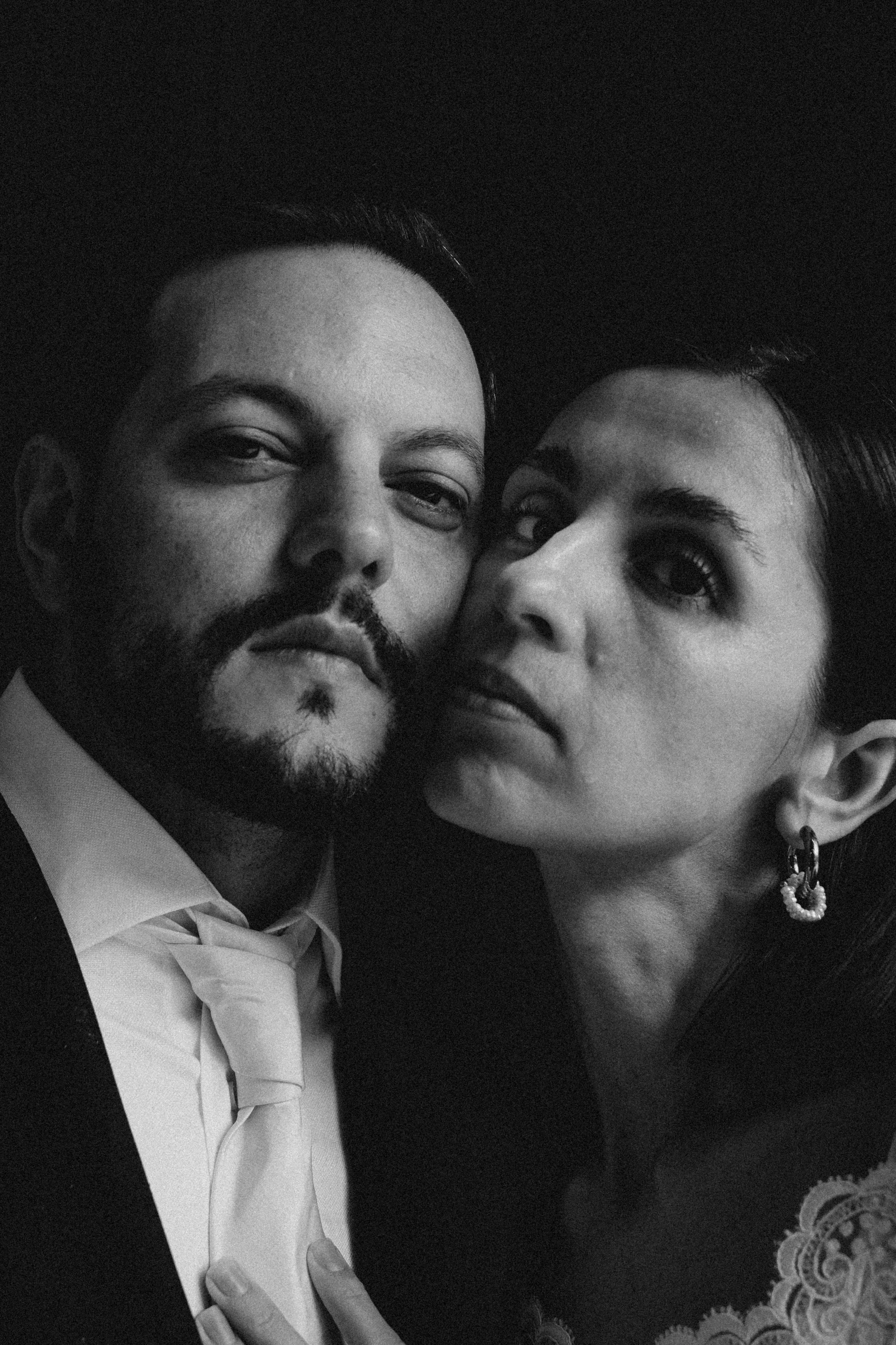 Una pareja tomados en una fotografía en blanco y negro, la mujer lleva un vestido con encaje y un pendiente grande, el hombre viste un esmoquin con pajarita.