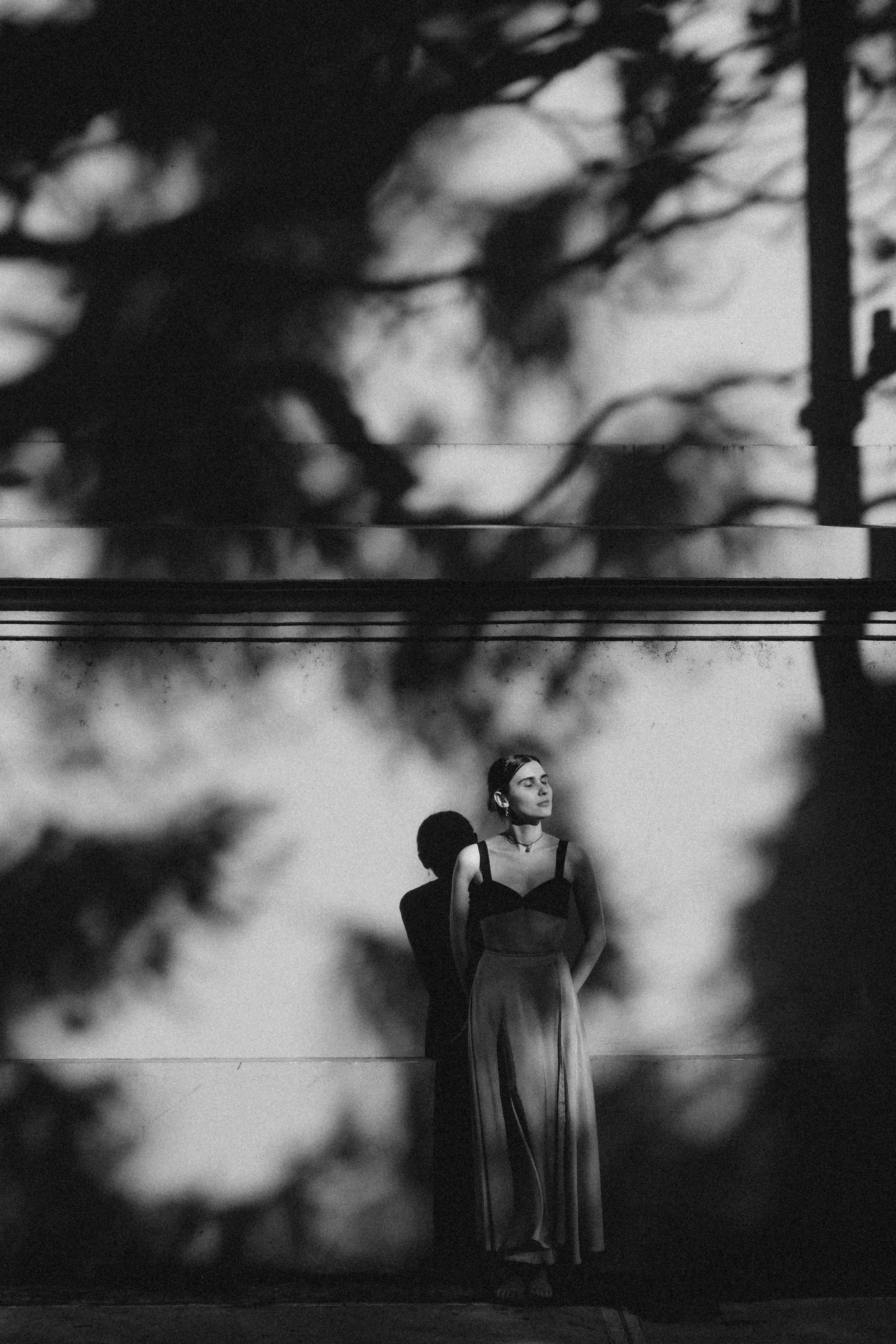 Mujer de cabello oscuro y ropa elegante de pie frente a un muro, con árboles nítidos y nubes en el fondo, en una foto en blanco y negro.