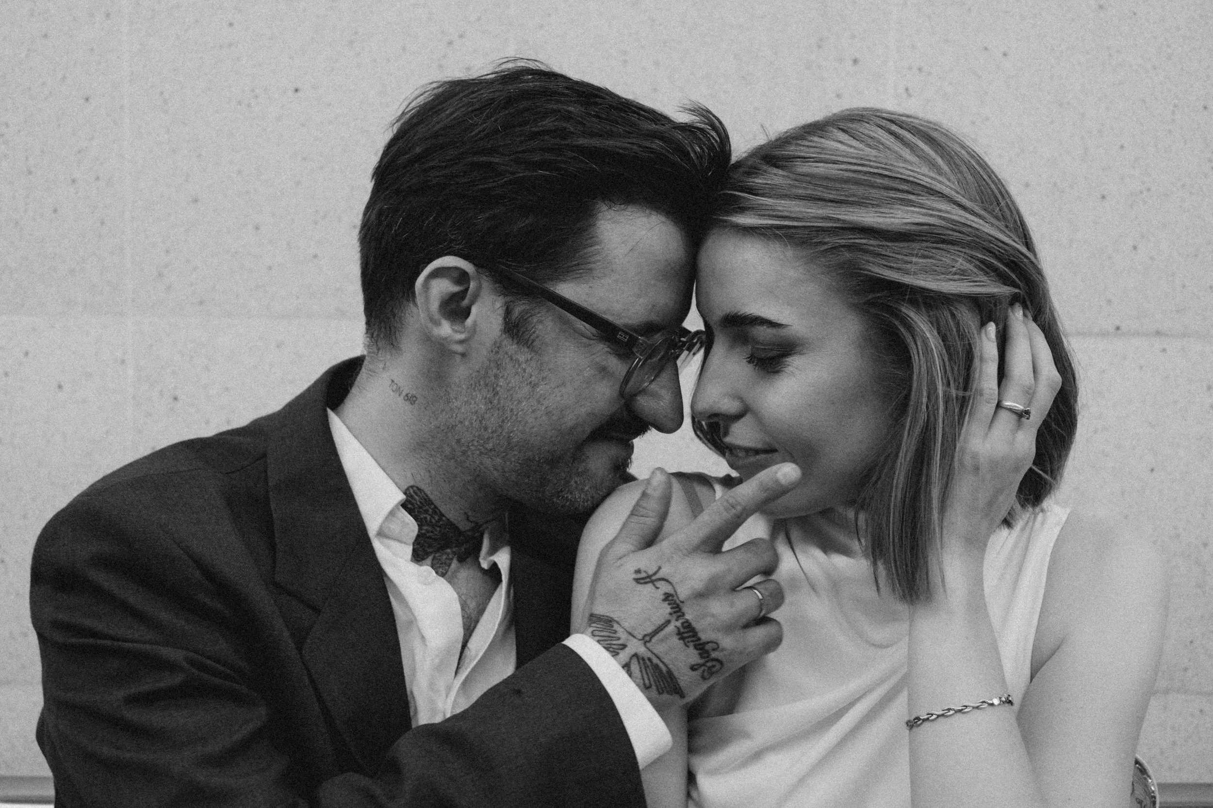 Una pareja se está acariciando y tocando la frente en una interacción íntima, en blanco y negro, con una pared de fondo.