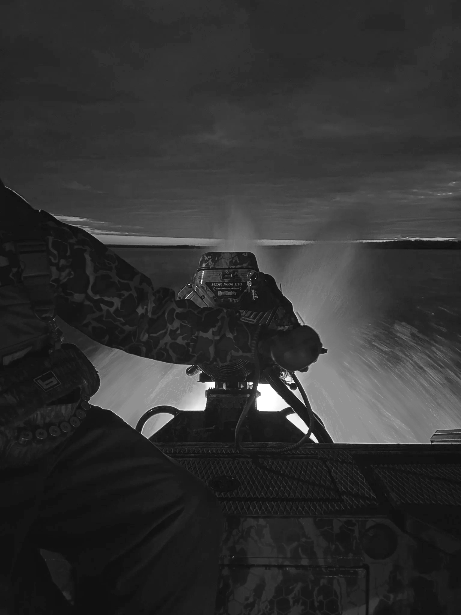 Black and white photo of a person operating a boat or watercraft, with spray of water and a distant horizon under a cloudy sky.
